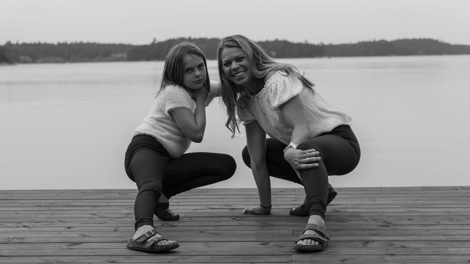 Two people crouch together on a wooden dock in a black and white photo.