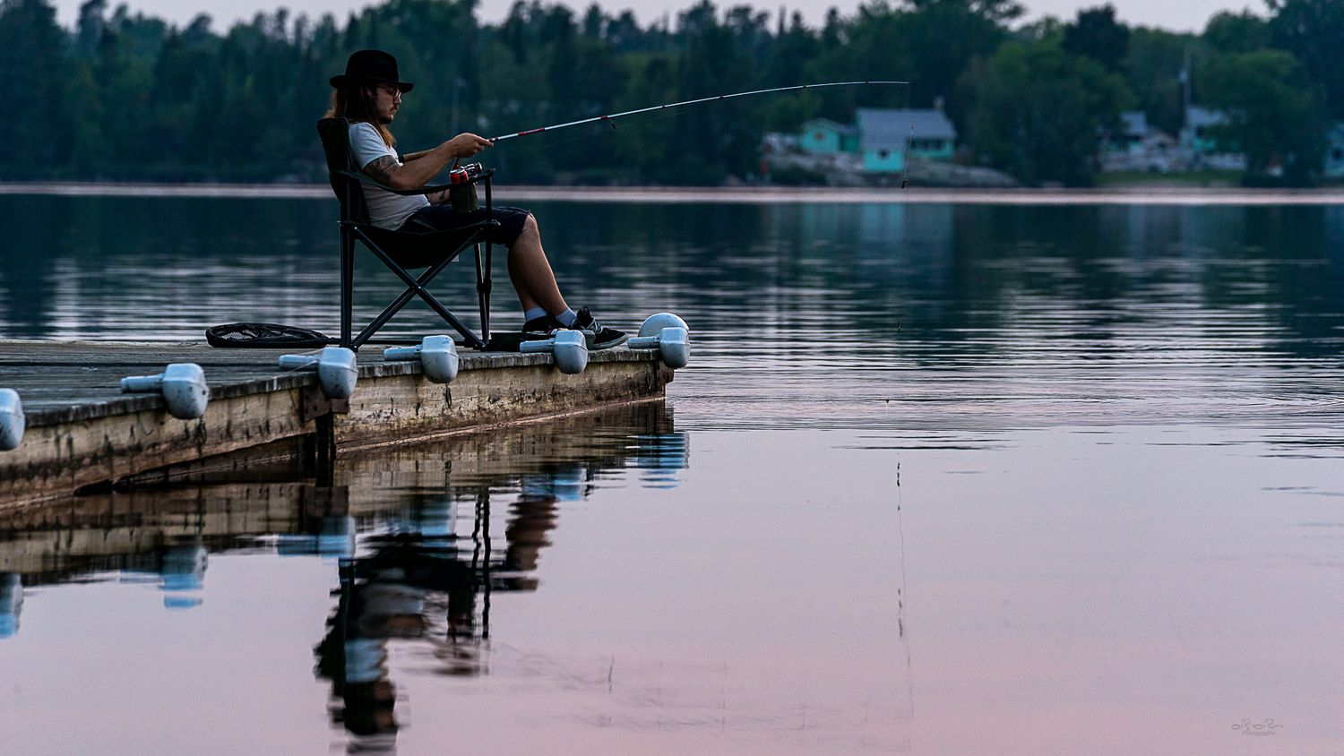 Silhouette of person fishing from dock at sunset with calm lake reflections.