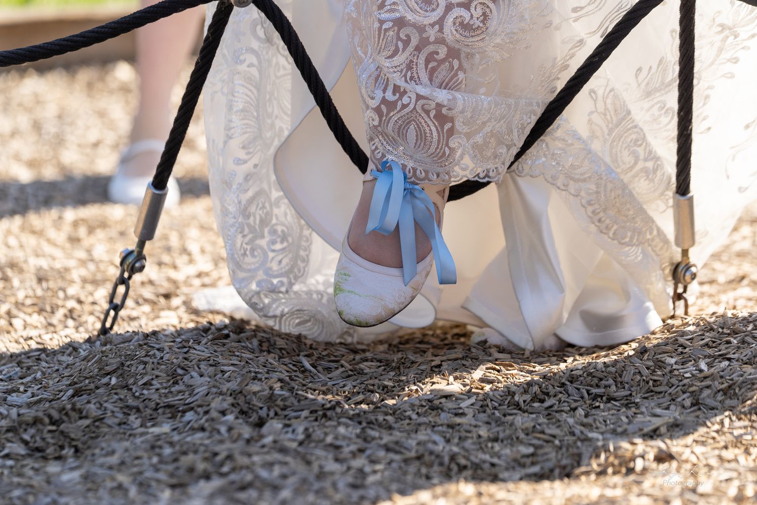 White wedding shoes peek out beneath a lace dress against gravel ground.