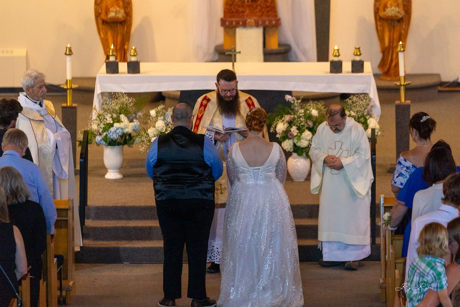 Wedding ceremony at church altar with white floral arrangements and clergy present.