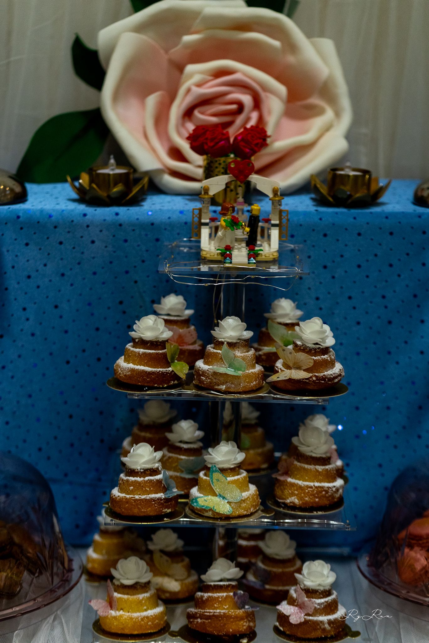 A tiered display of frosted mini cakes arranged on a stand with a pink rose and gold candle holders in the background.