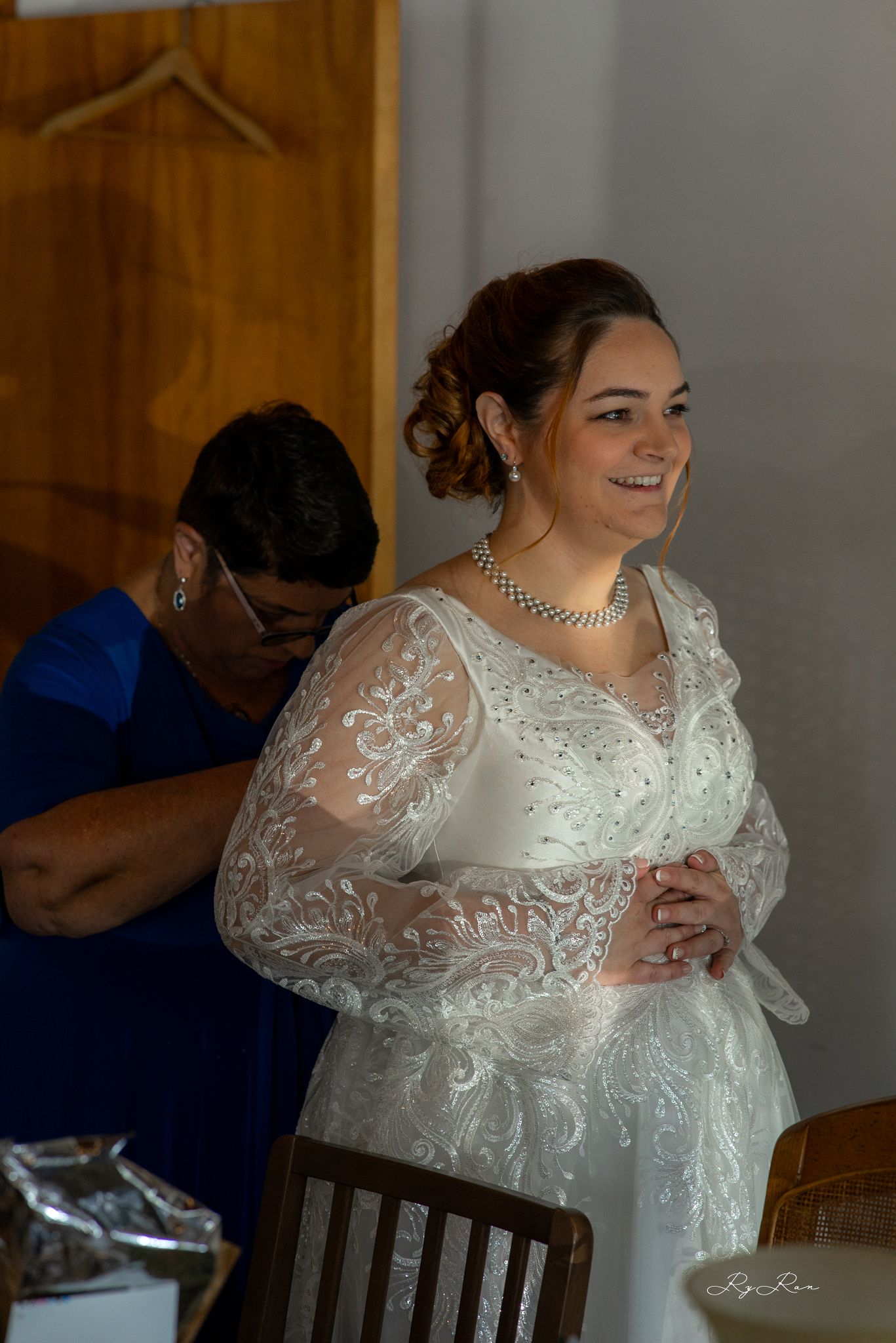 A bride wearing an elegant lace wedding gown with sheer sleeves smiles while getting ready.