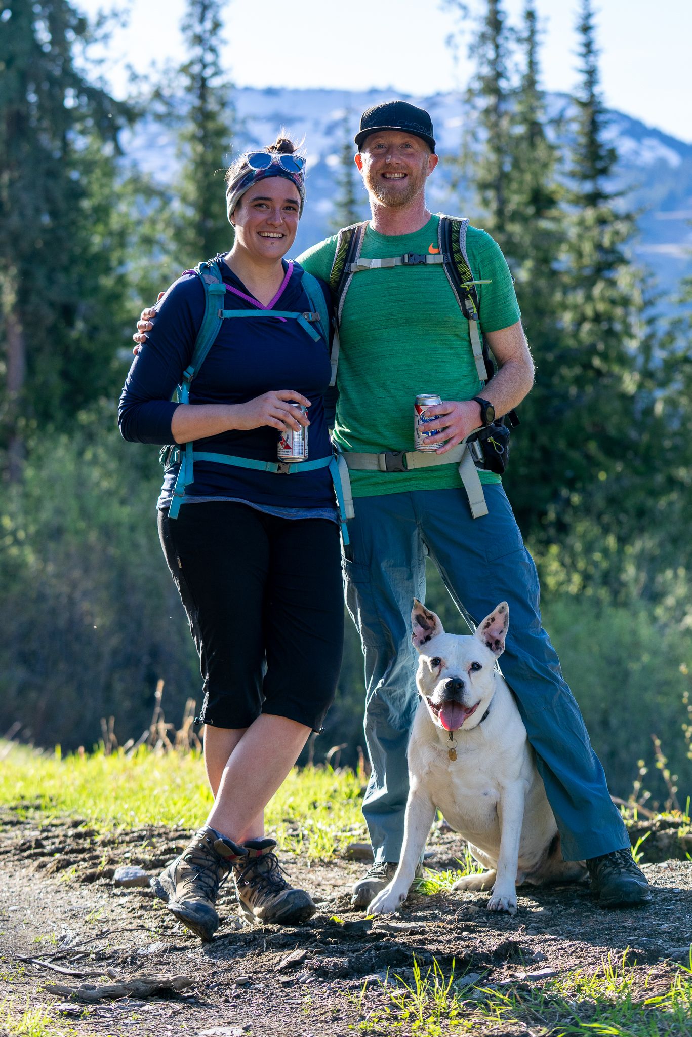 Hikers with backpacks and their happy white dog pause on mountain trail.