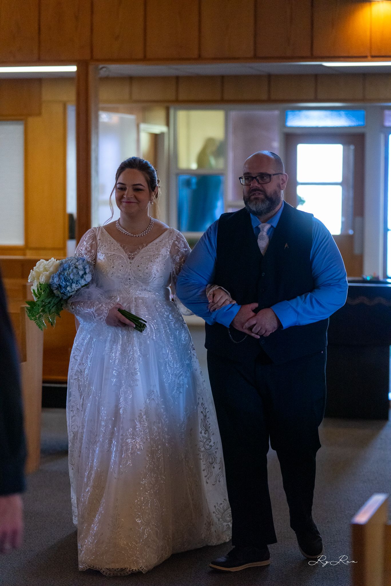 A bride in a white lace gown walks down the aisle with an escort.