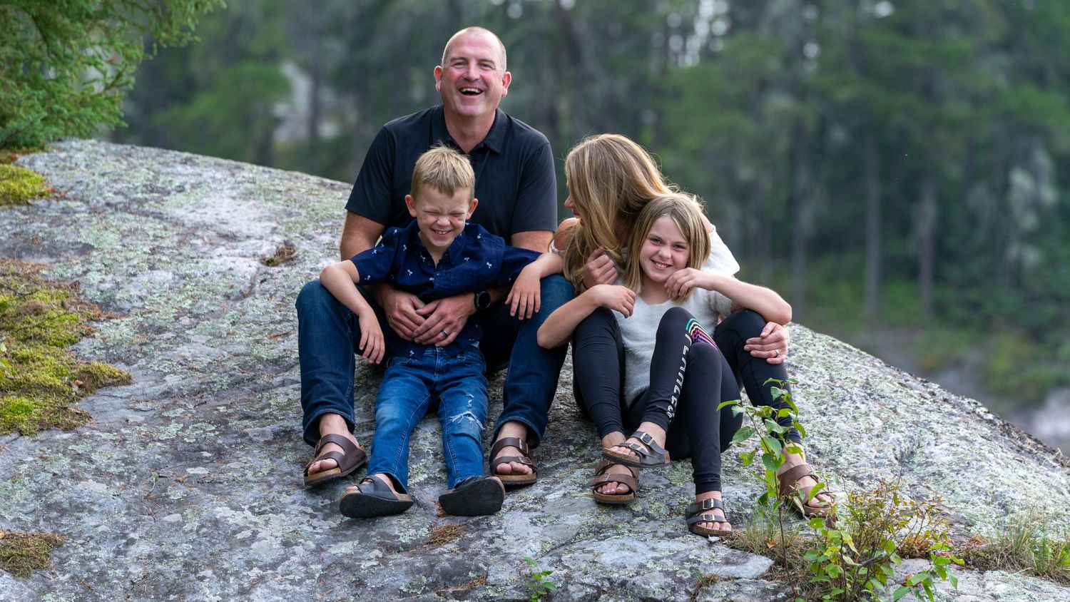 Family of four sitting together and smiling on large rock formation outdoors.