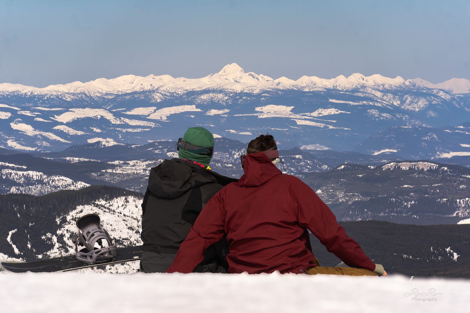 Two people sitting on snowy mountain summit overlooking snow-capped peaks against blue sky.