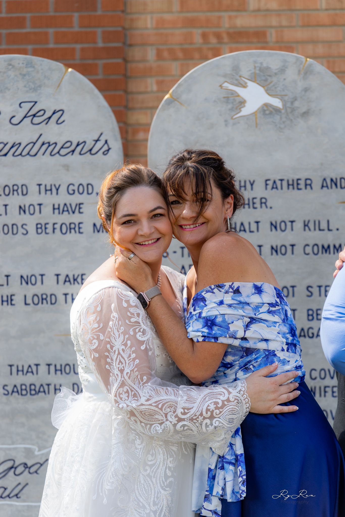Two people share a joyful embrace in front of stone tablets, one in a wedding dress and one in blue.