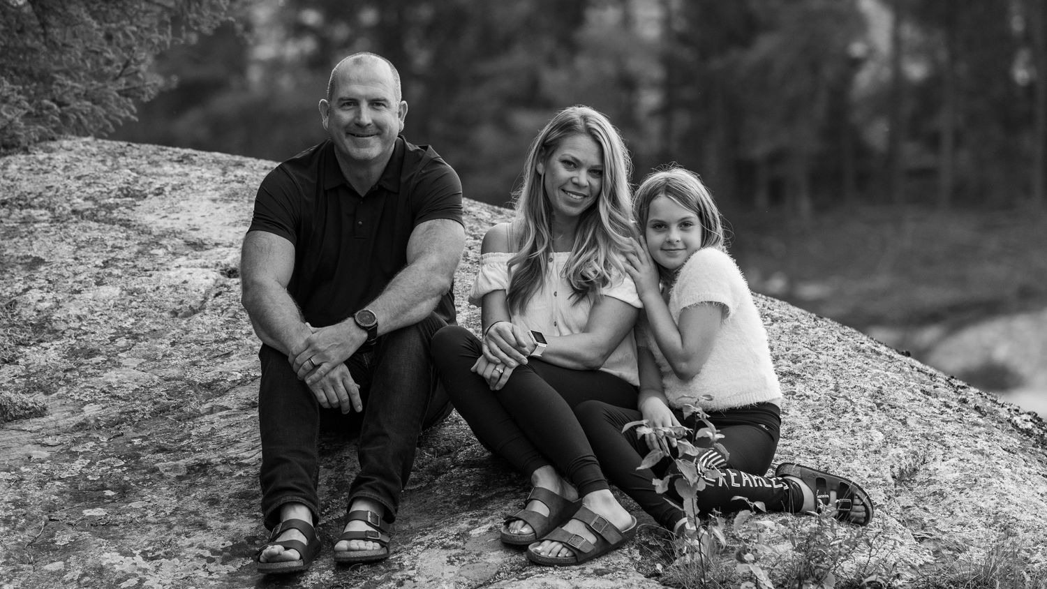Black and white family portrait of three people sitting together on rocky surface outdoors.