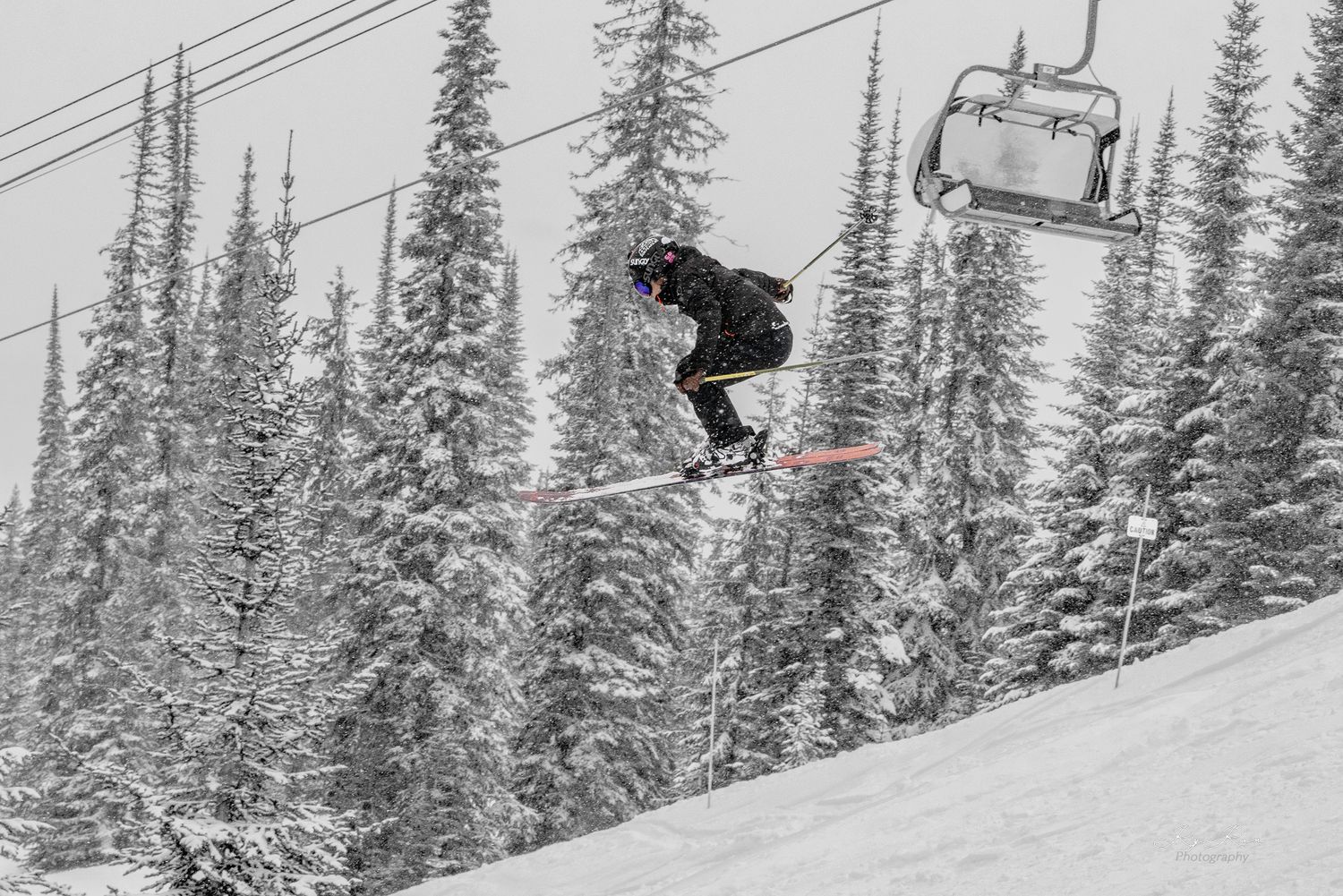 A snowboarder performing a jump with a ski lift and snow-covered evergreen trees in the background.