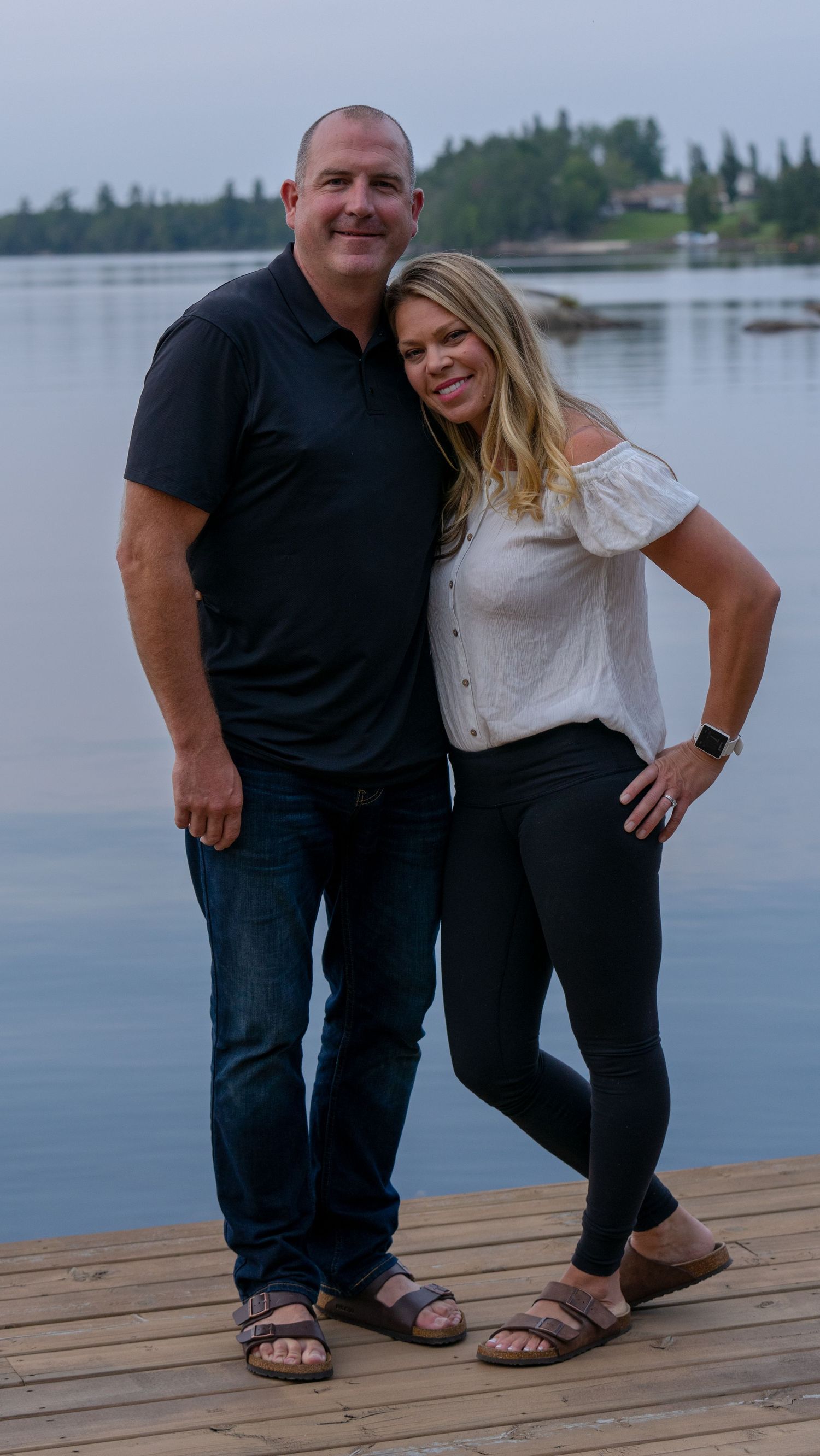 A couple stands together on a wooden dock with a peaceful lake and forest backdrop.