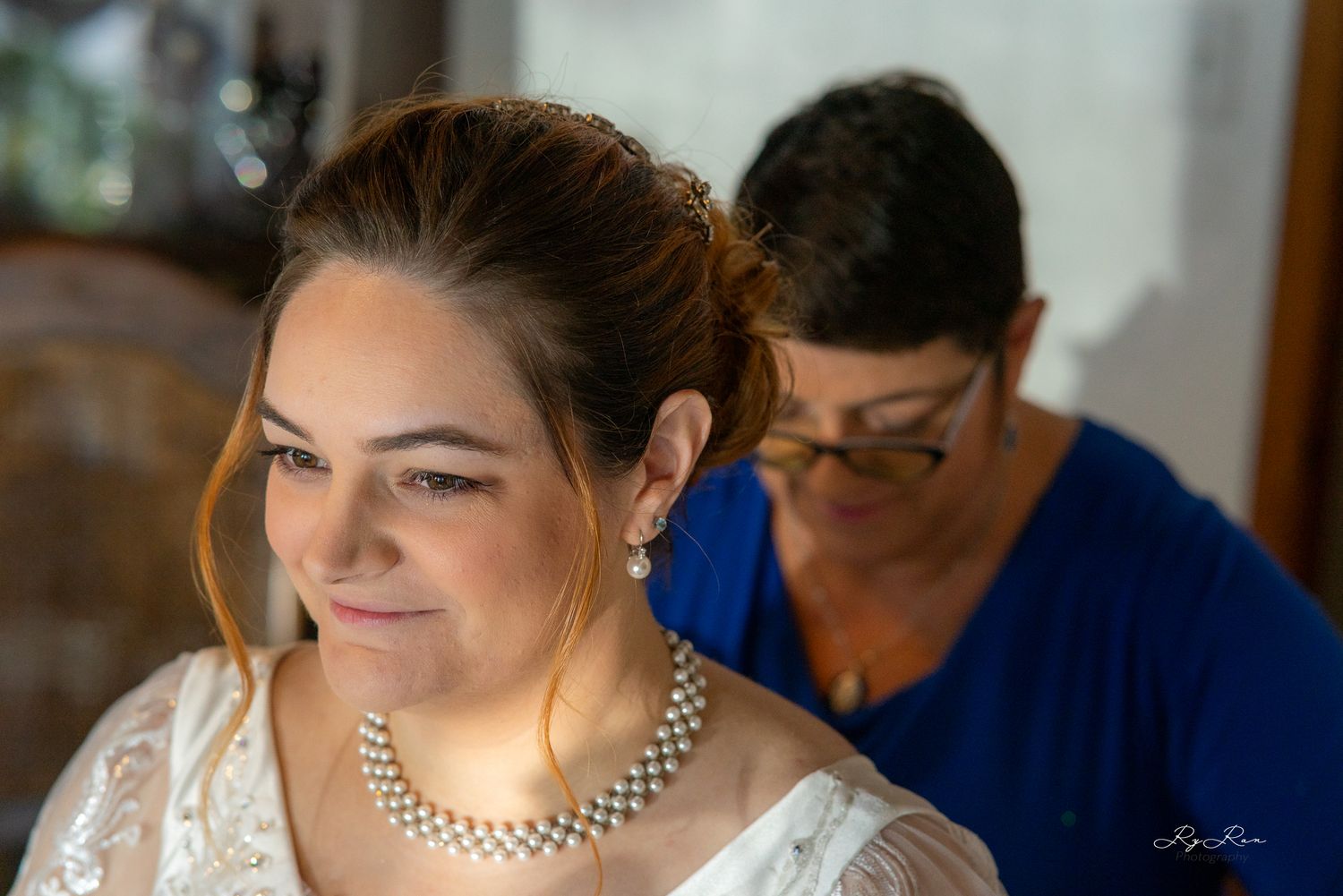 A smiling bride in white lace dress and pearl necklace during wedding preparations.