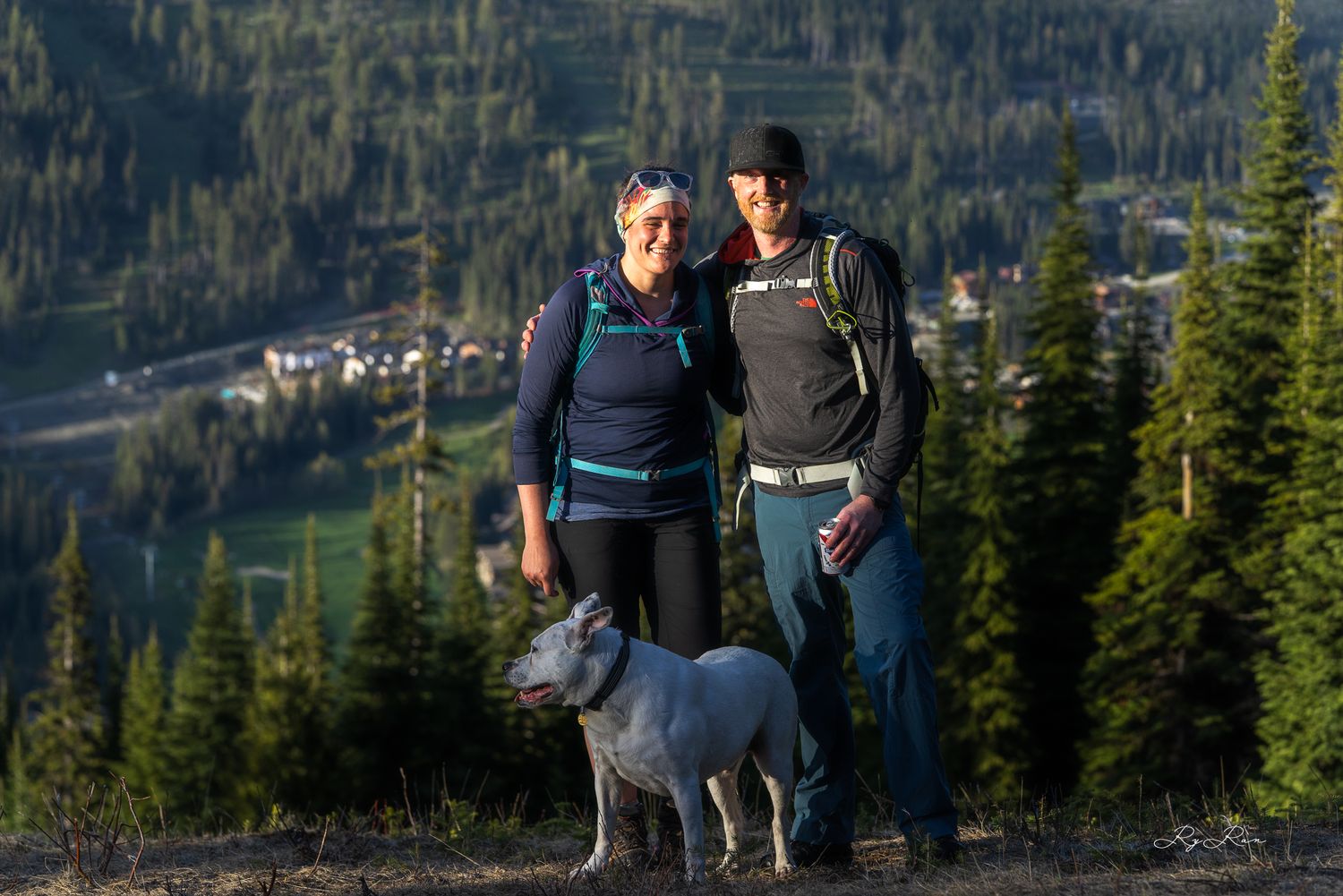 Two hikers and their dog pose together on mountain overlook with valley view.