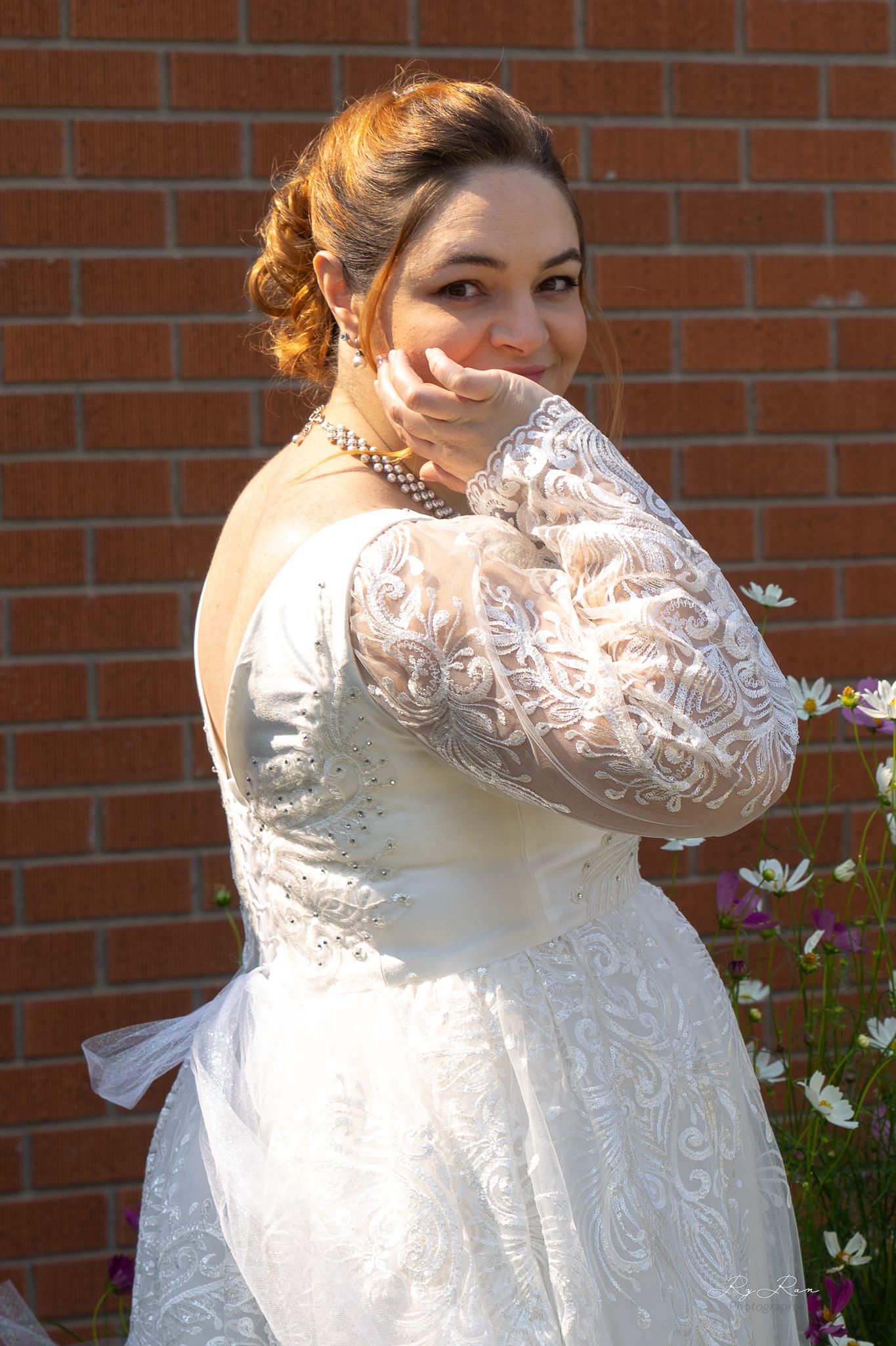 Elegant lace wedding gown with long sleeves and intricate detailing shown against brick wall and flowers.