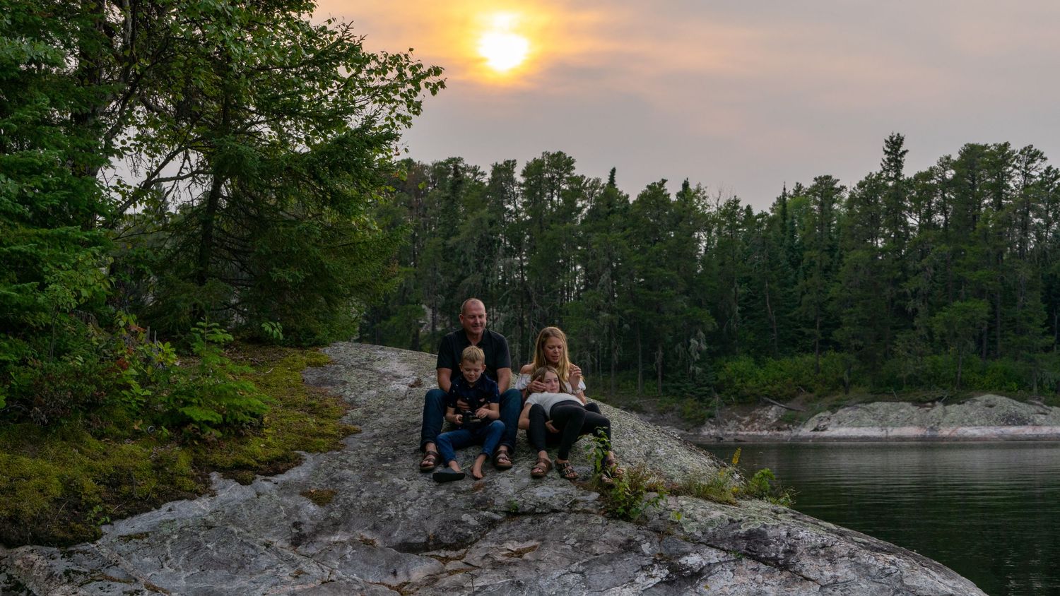 Family posing on a rocky cliff at sunset with a lake and pine forest in the background.