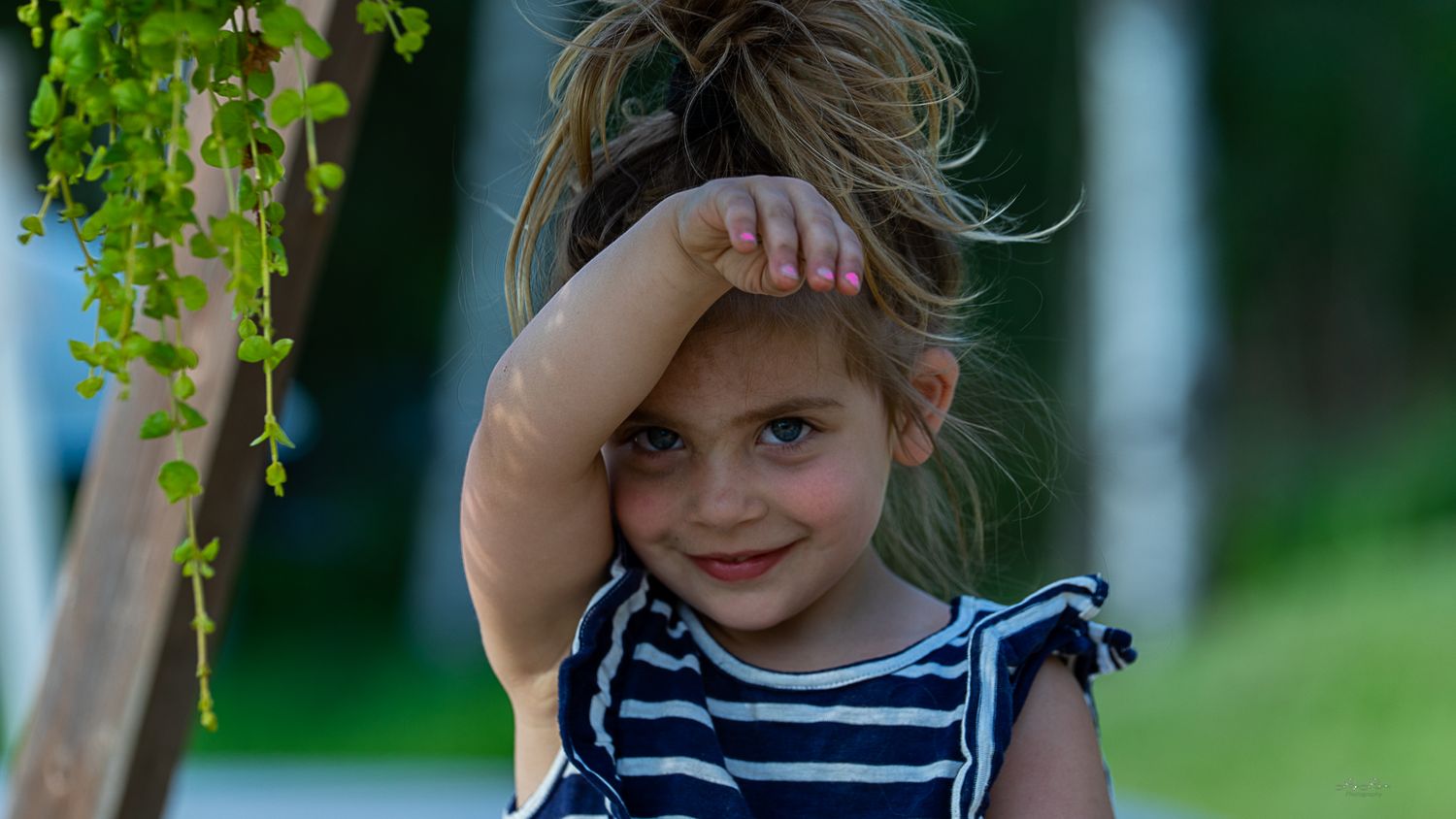 A young child in a striped dress shielding eyes from sun with greenery in background.