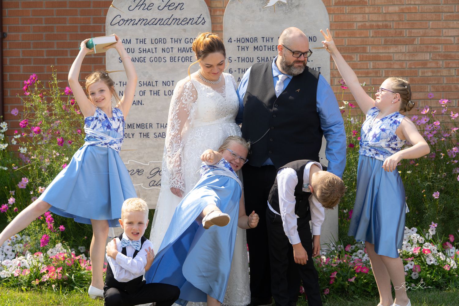 Playful wedding party poses for fun group photo in blue and white outfits by garden.