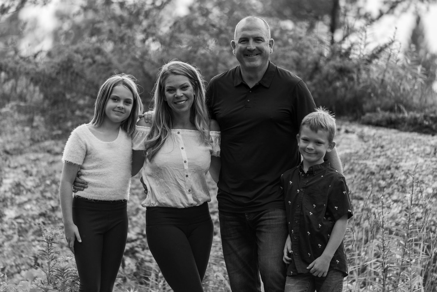 Family of four posing for black and white portrait against natural outdoor setting.