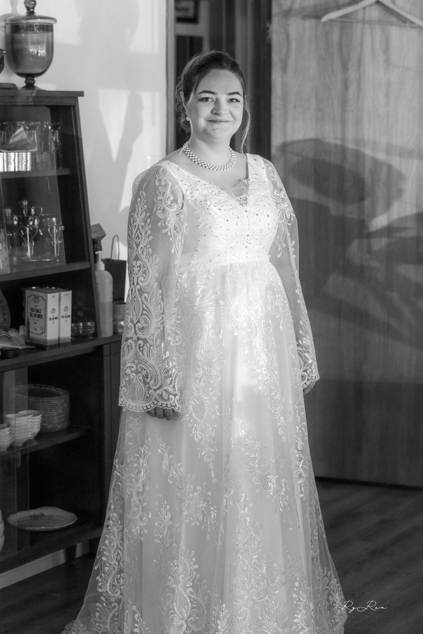 A bride poses in a flowing white lace wedding gown with long sleeves next to a wooden cabinet.