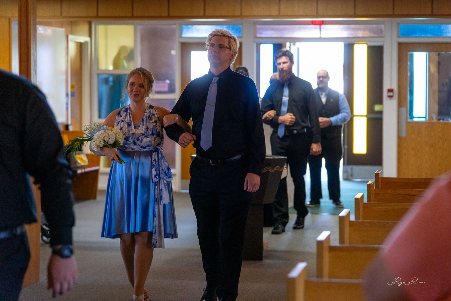 Wedding processional in a church hallway with attendants in blue dresses.
