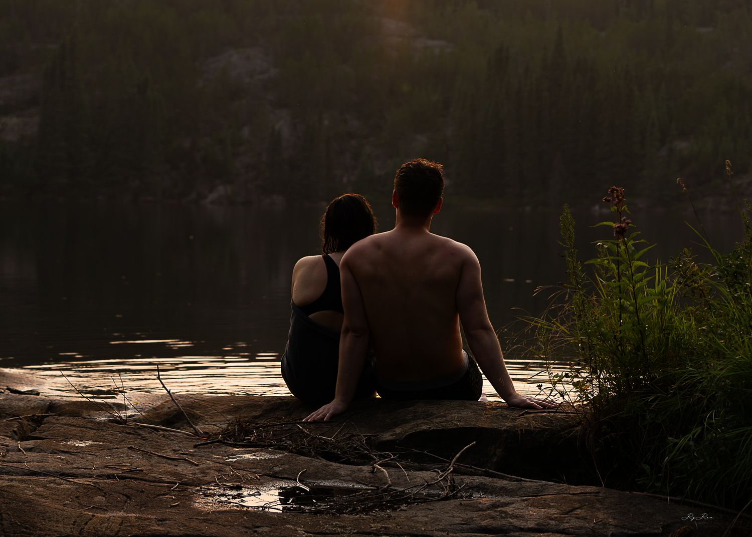 Silhouette of two people sitting together by a lake at sunset enjoying a peaceful moment.