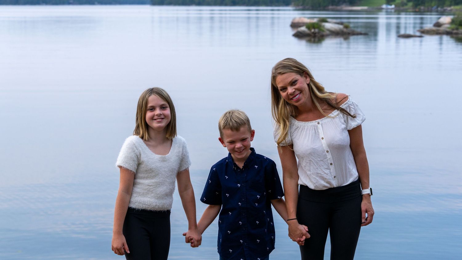 Three people hold hands while standing together on a dock by a calm lake.