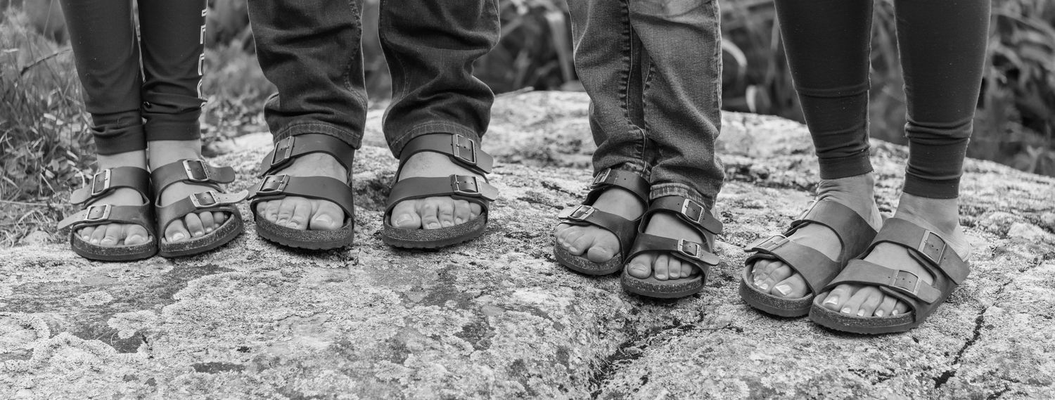 Black and white close-up of four pairs of feet wearing sandals standing on rock surface.