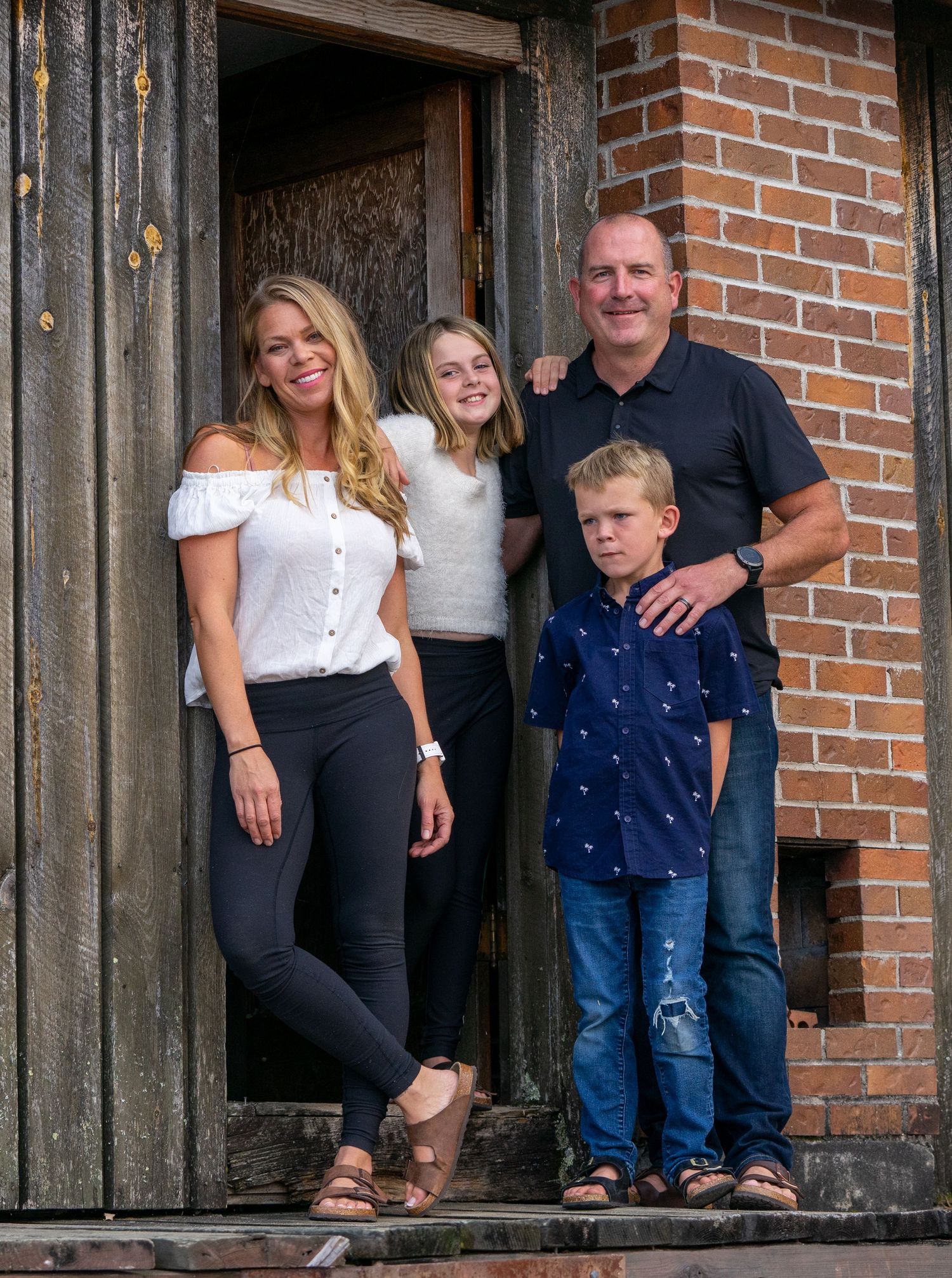 Family of four poses together on wooden porch steps with rustic backdrop.