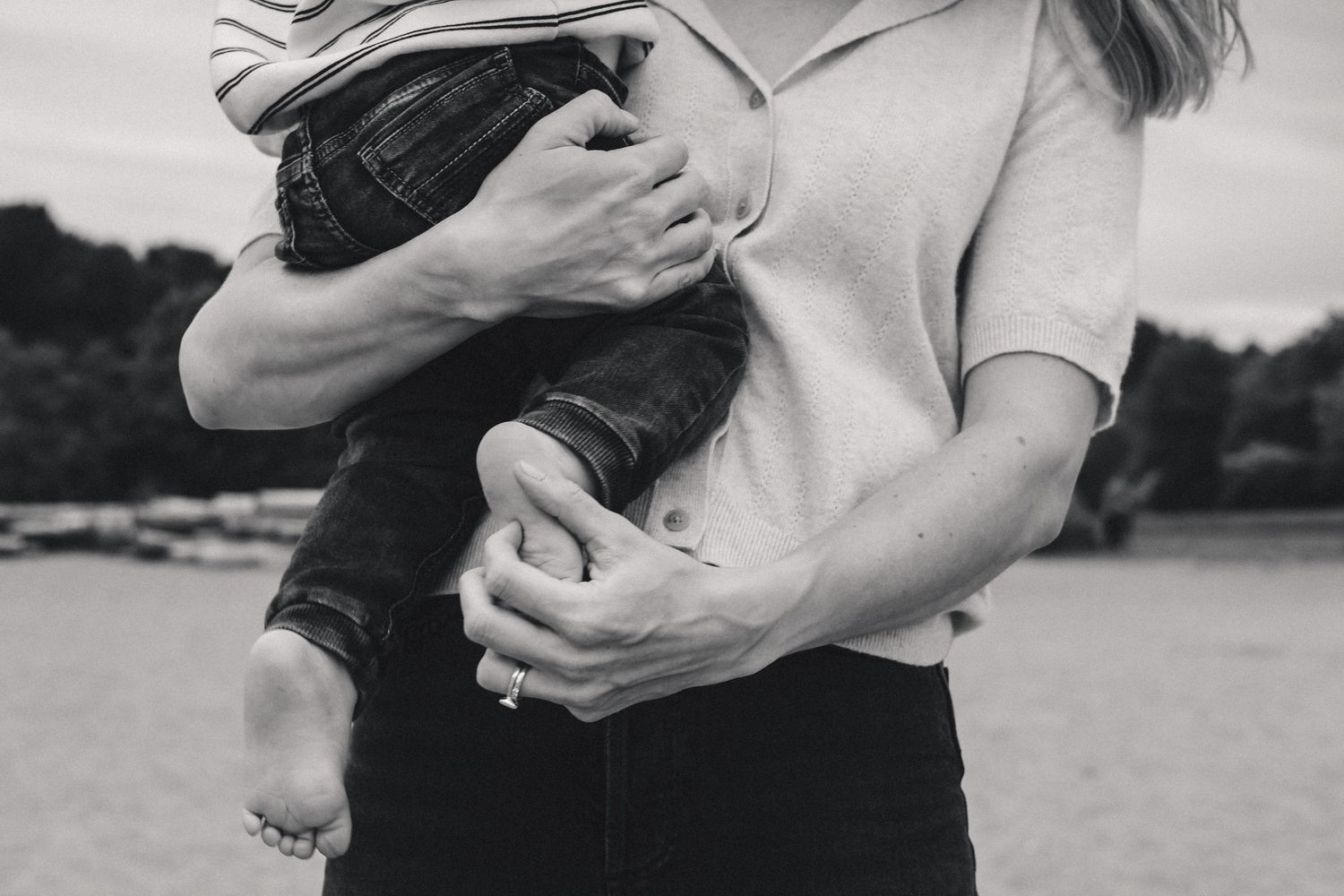 A parent holds a young child close against a waterfront background in black and white.
