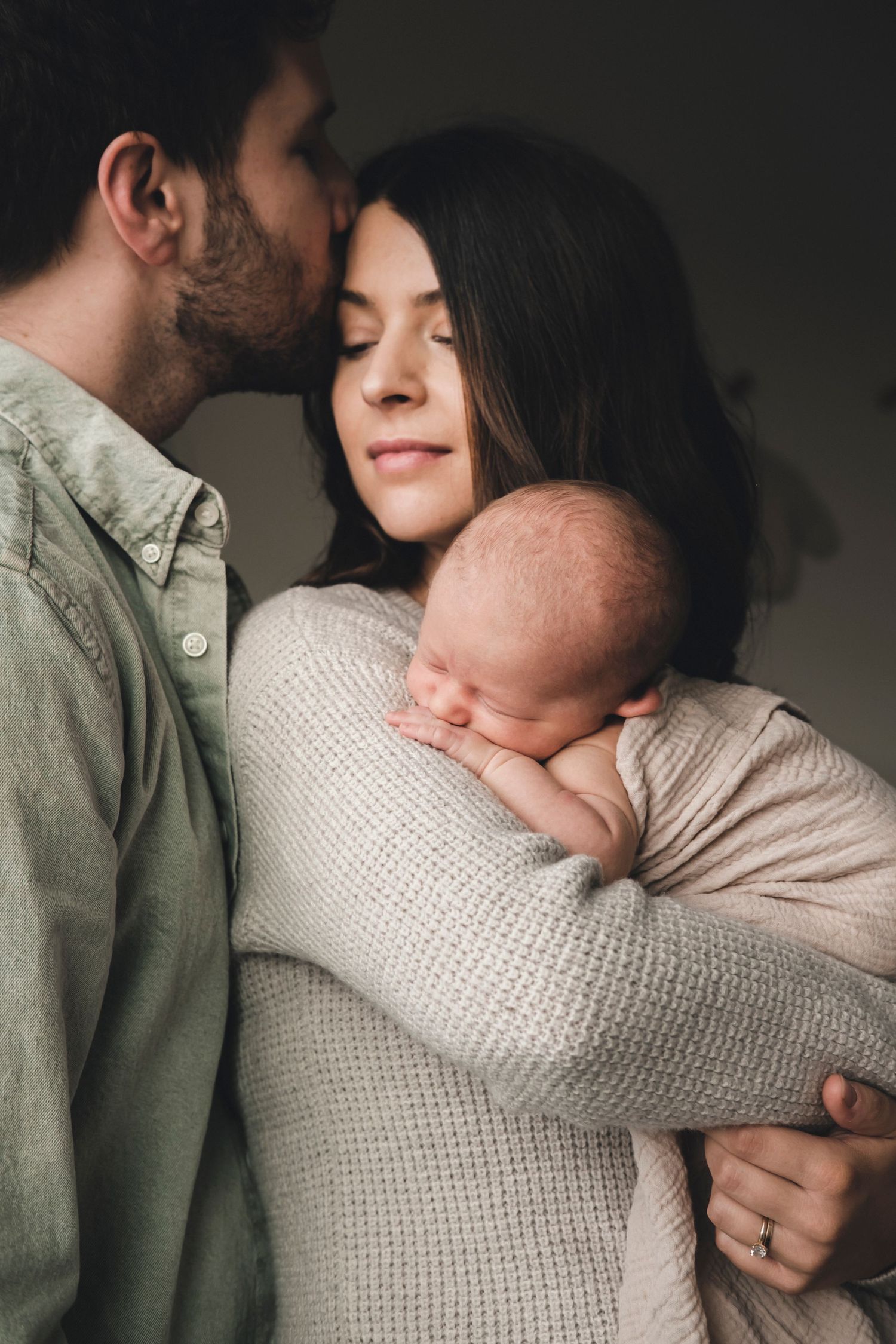 Parents share tender moment with sleeping newborn baby wrapped in knit blanket.