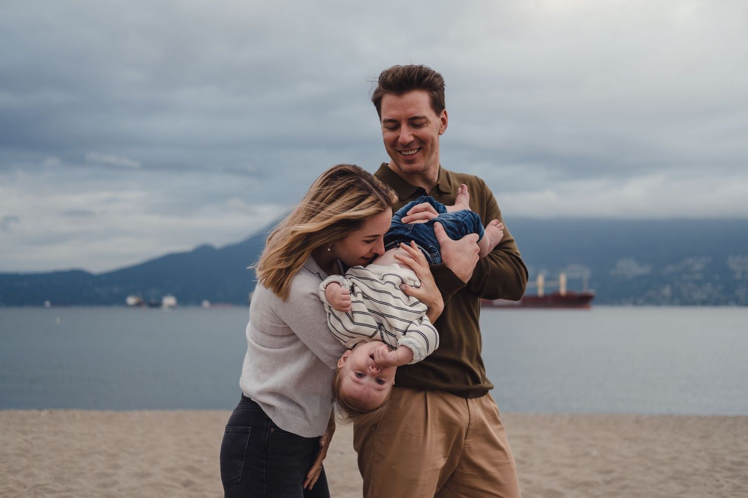 Parents share playful moment with baby against scenic mountain ocean backdrop.