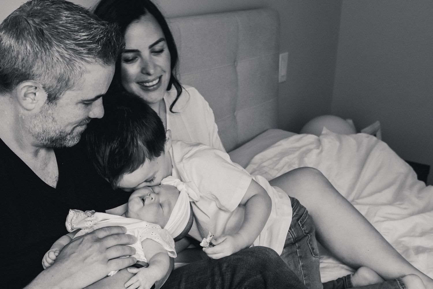 Intimate black and white family moment as parents share tender interaction with newborn baby in bed.
