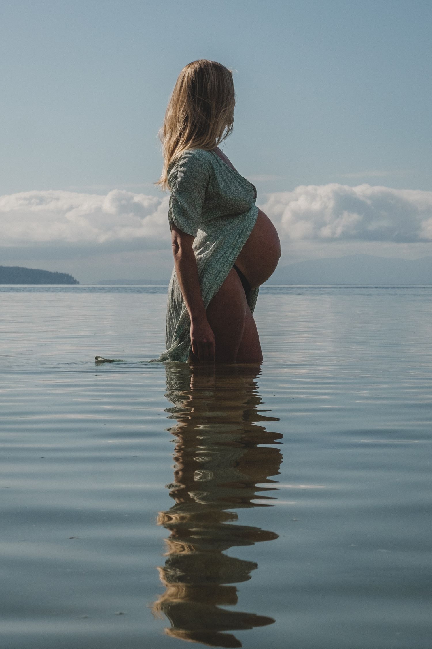 Atmospheric photo of a woman in a dress standing in calm reflective water with mountains and clouds in the distance.