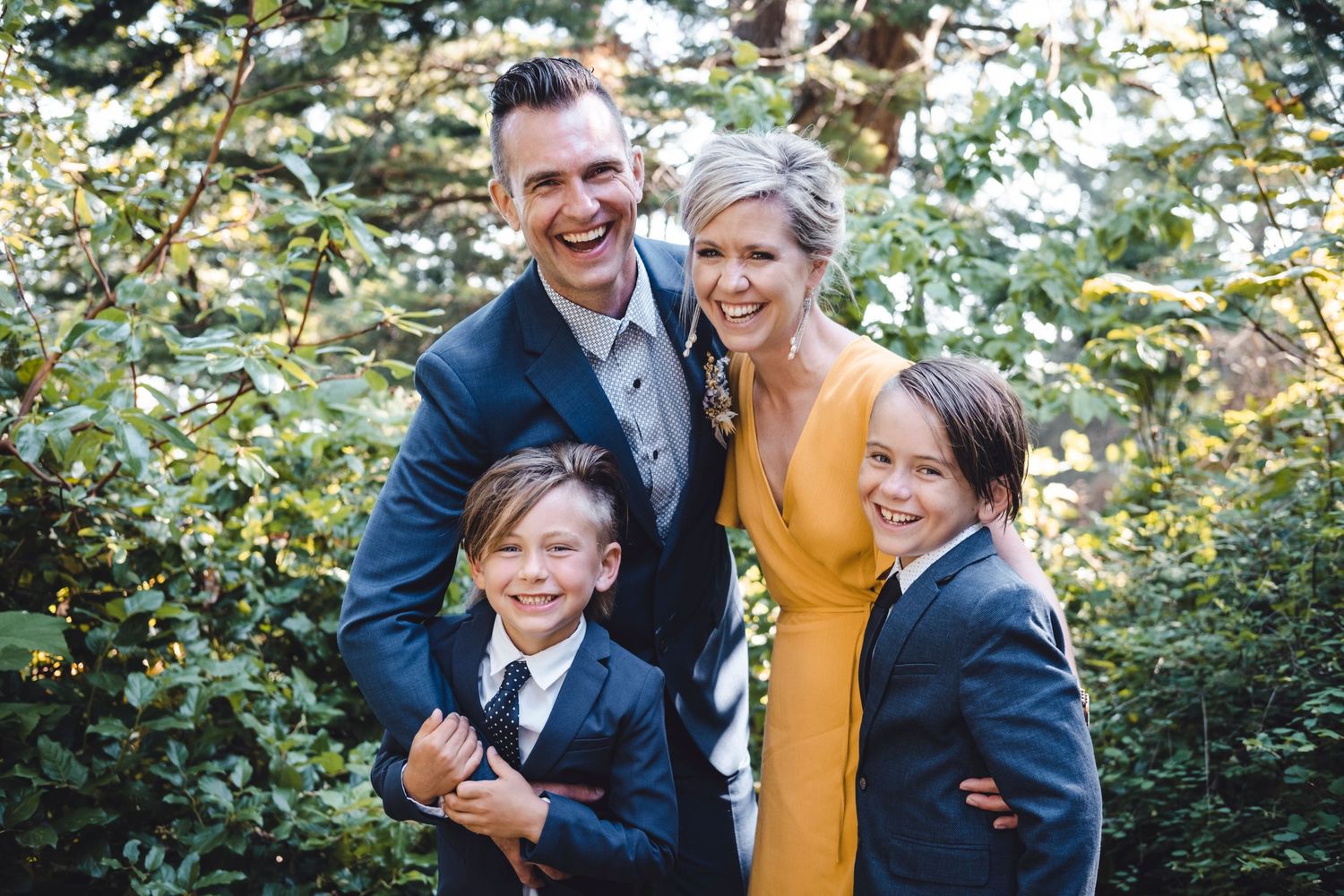 Family of four poses together outdoors in natural setting with greenery background.