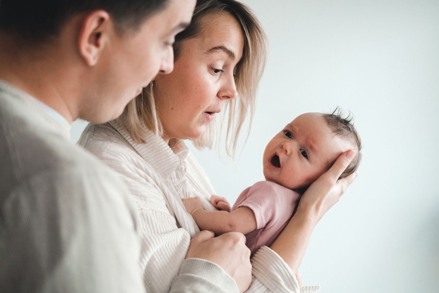 Parents lovingly hold and gaze at their newborn baby in a tender moment against a light background.