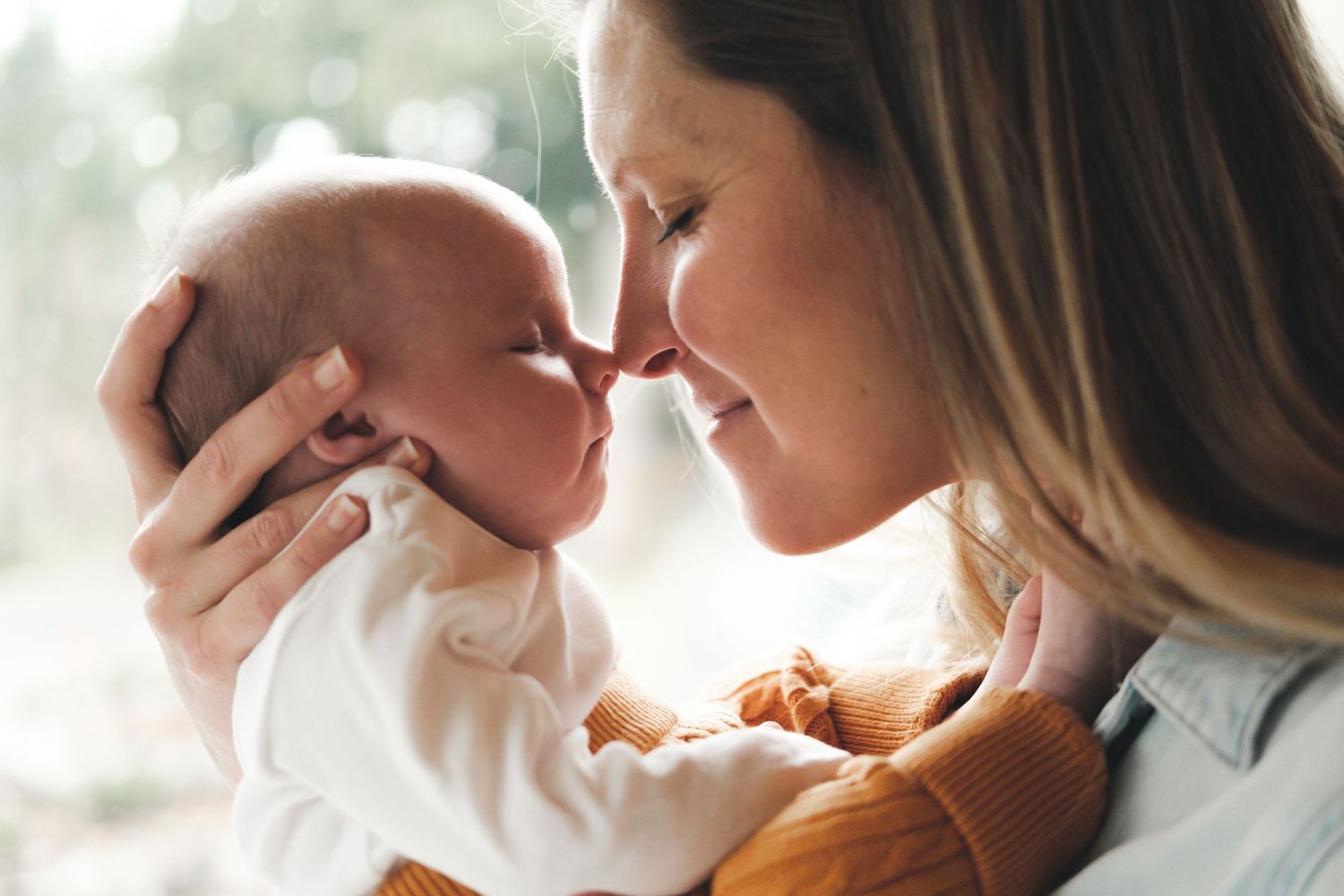 Mother and newborn share intimate nose-to-nose moment in soft window light.