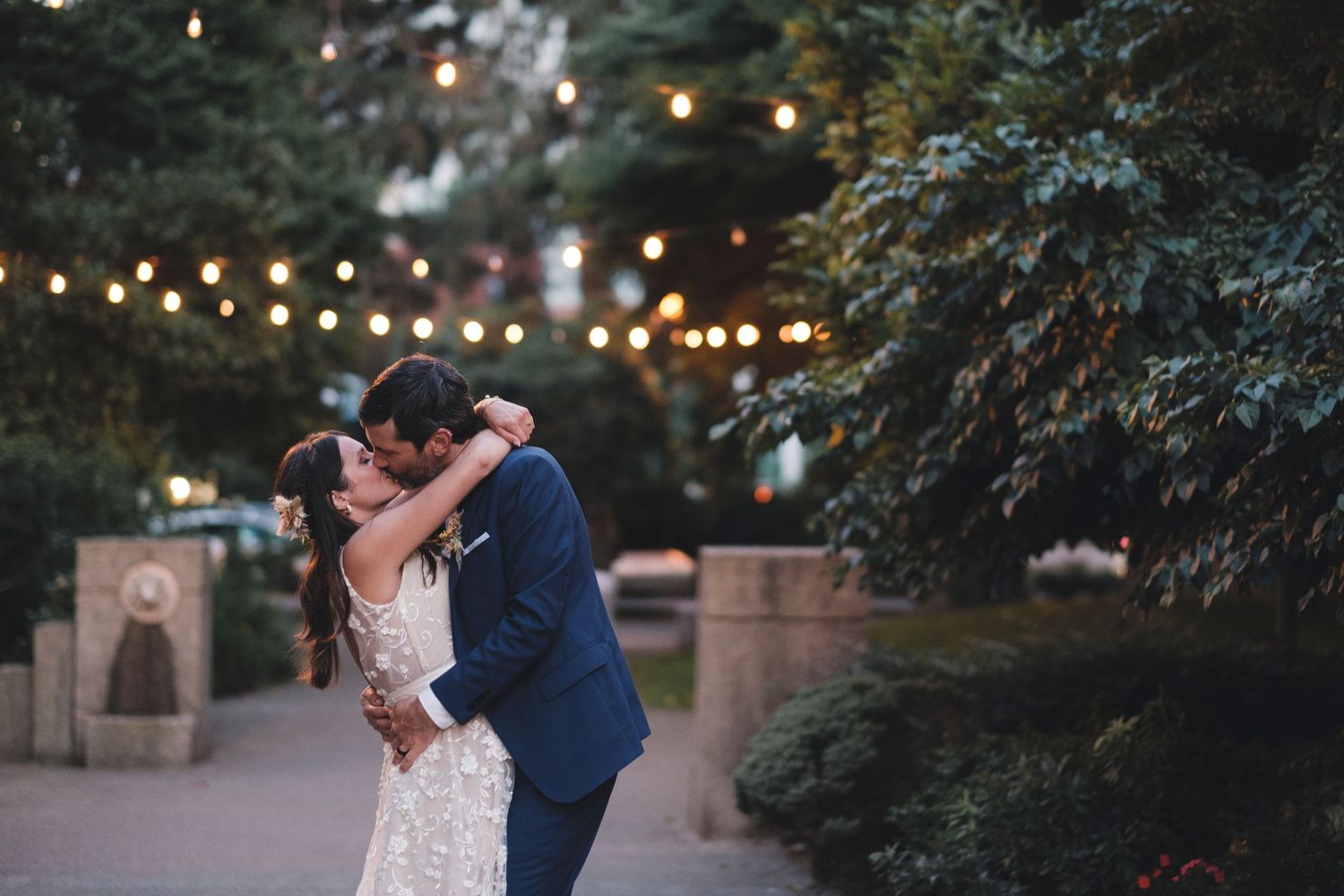 Couple shares romantic embrace under string lights during evening wedding celebration.