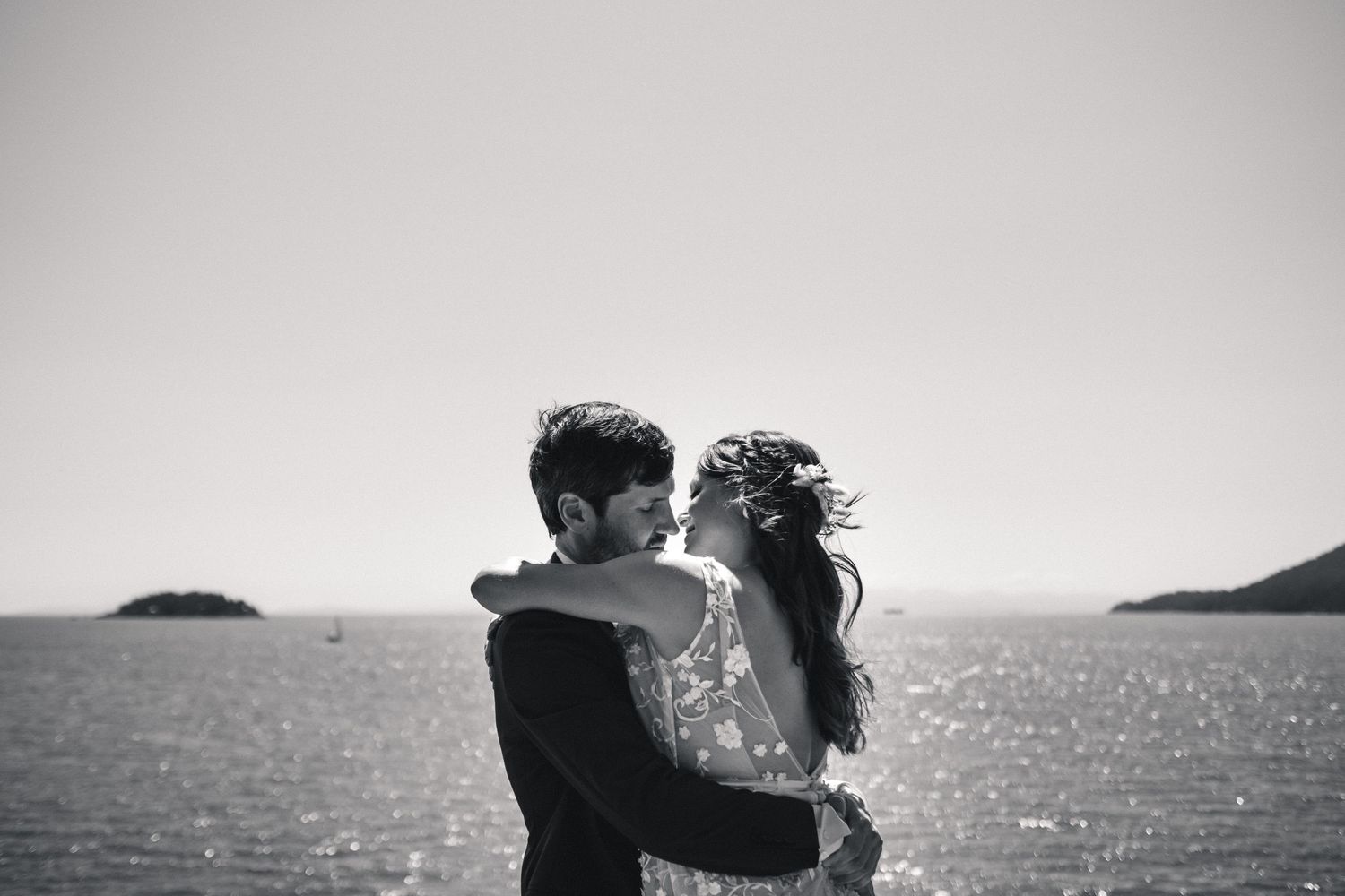 Black and white silhouette of couple embracing by ocean with islands in background.