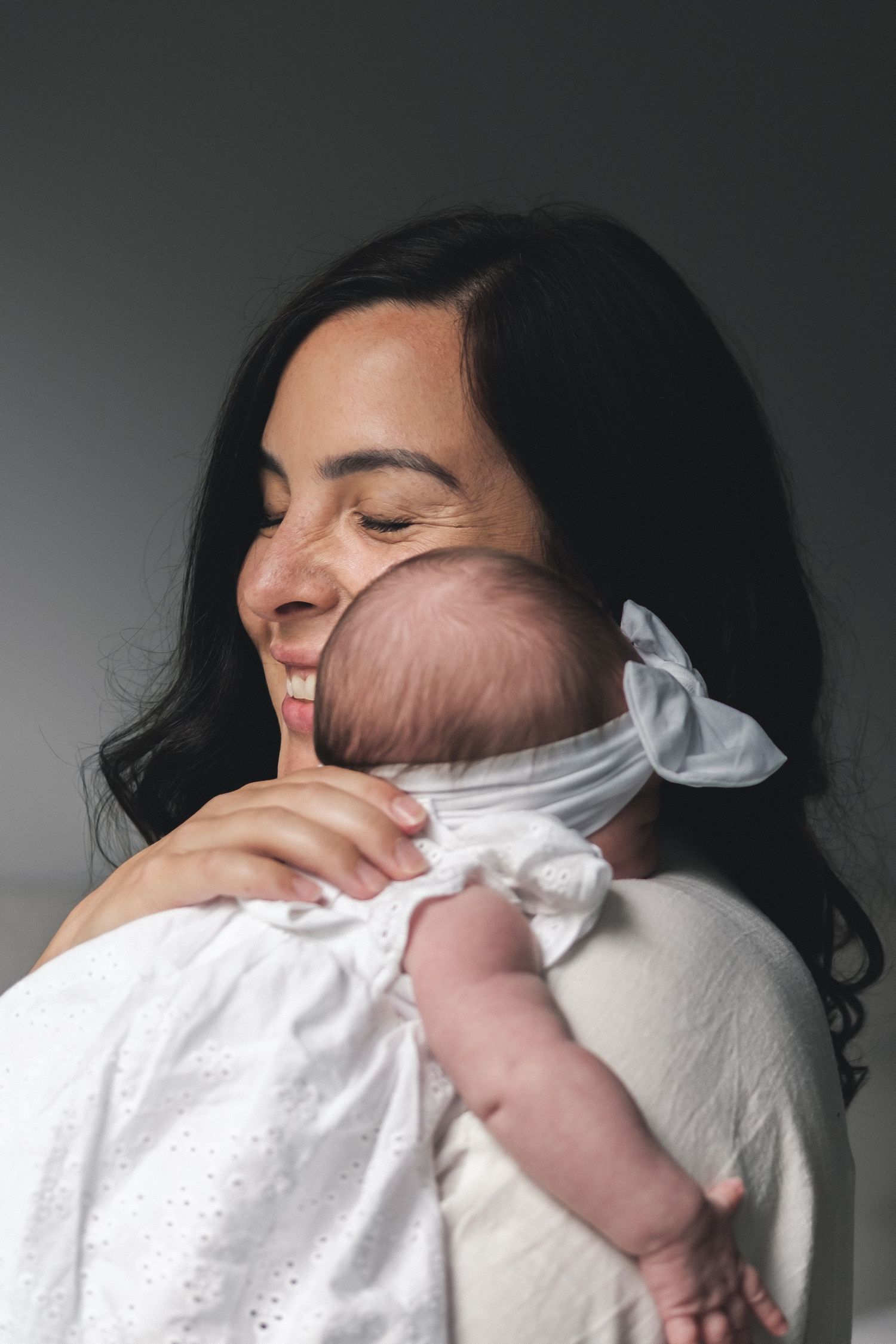 Mother in dark hair embraces infant wearing white clothing in emotional portrait against grey background.