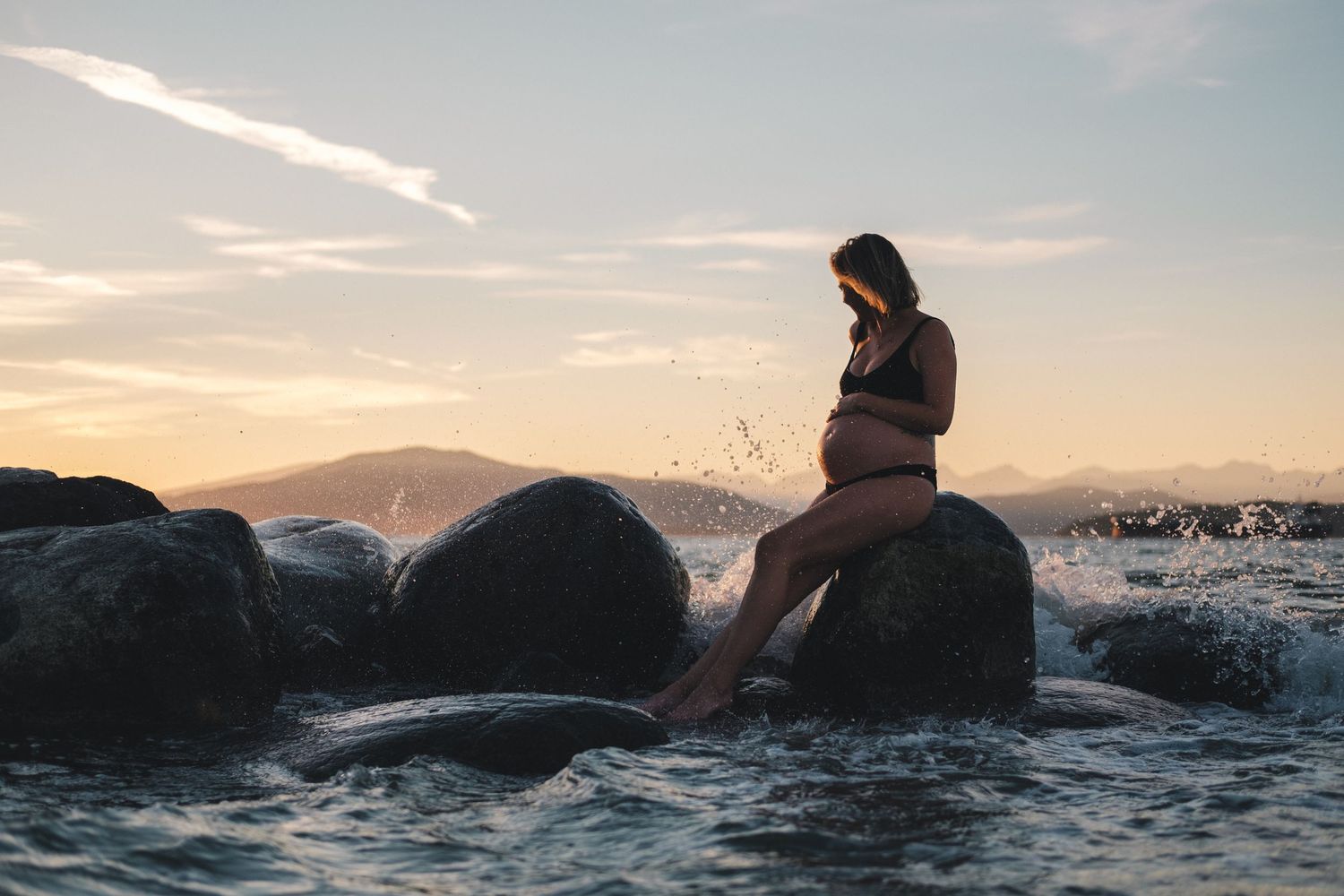 Silhouette of person sitting on rocks by ocean during golden sunset.