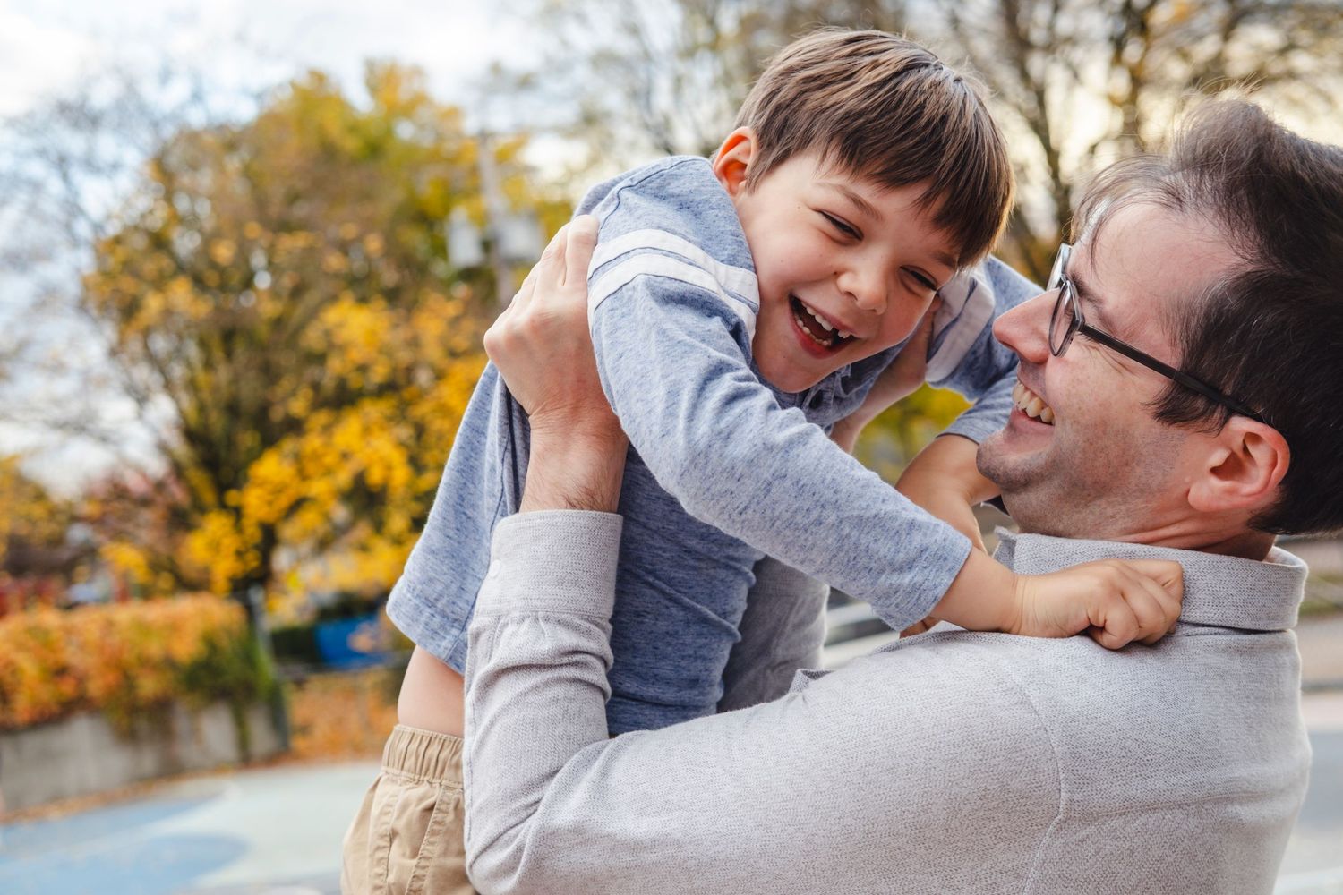 A playful moment between parent and child sharing laughter outdoors with autumn foliage in the background.
