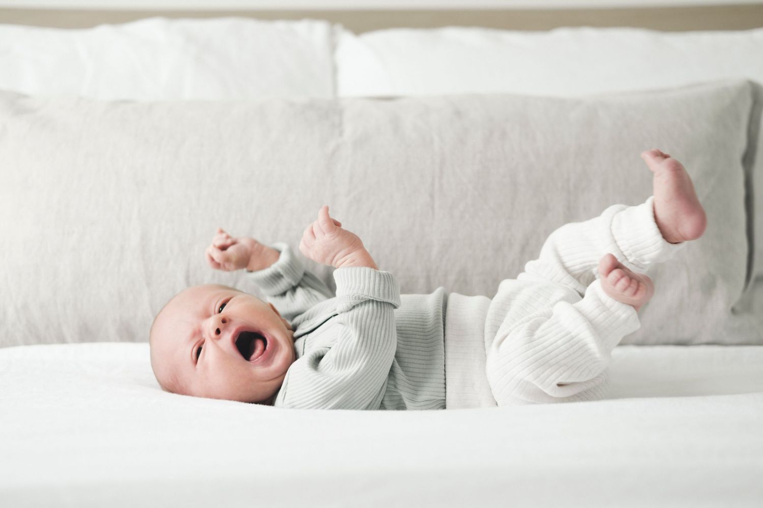 Laughing baby in white onesie lying on white bedding with arms and legs up.