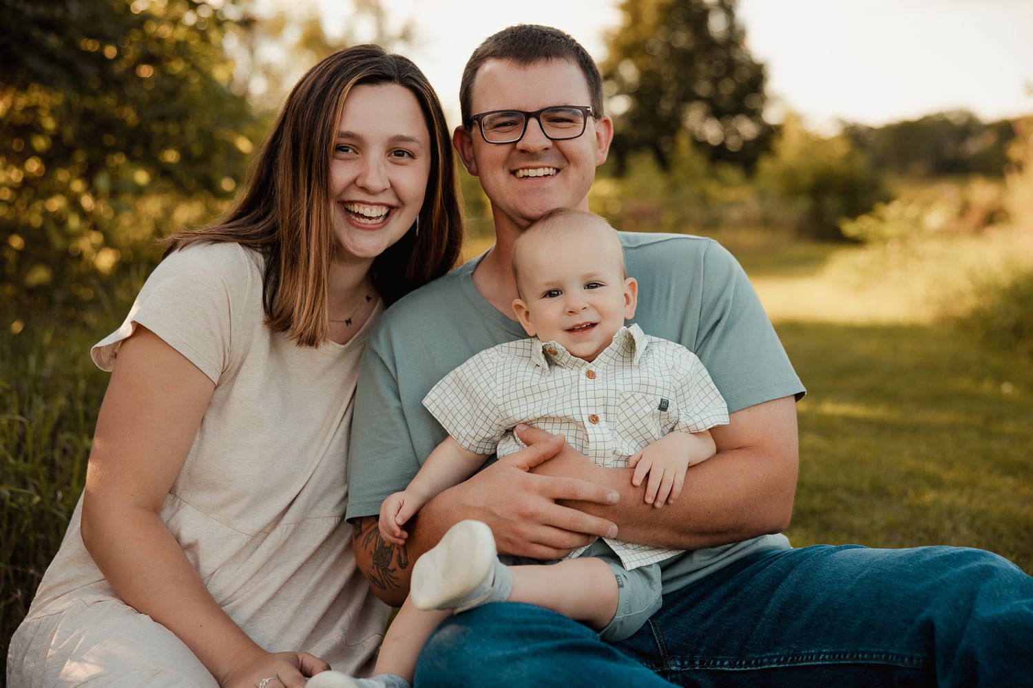Happy young family with smiling parents and baby boy sitting outdoors in a sunny green park setting.
