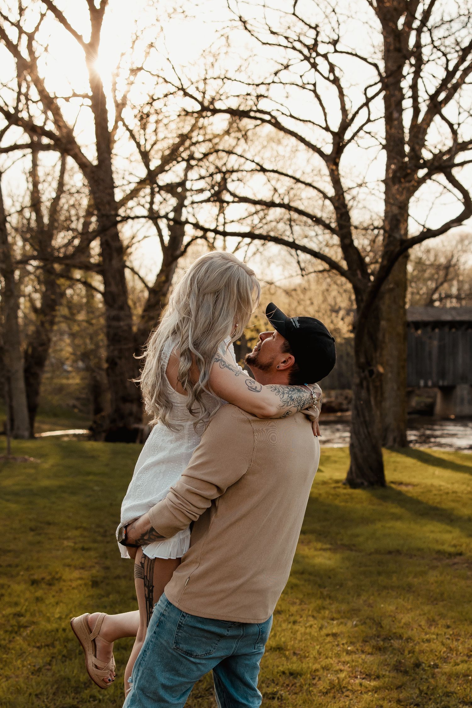 A man lifts a woman in a romantic embrace outdoors, surrounded by bare trees with golden sunset light filtering through.