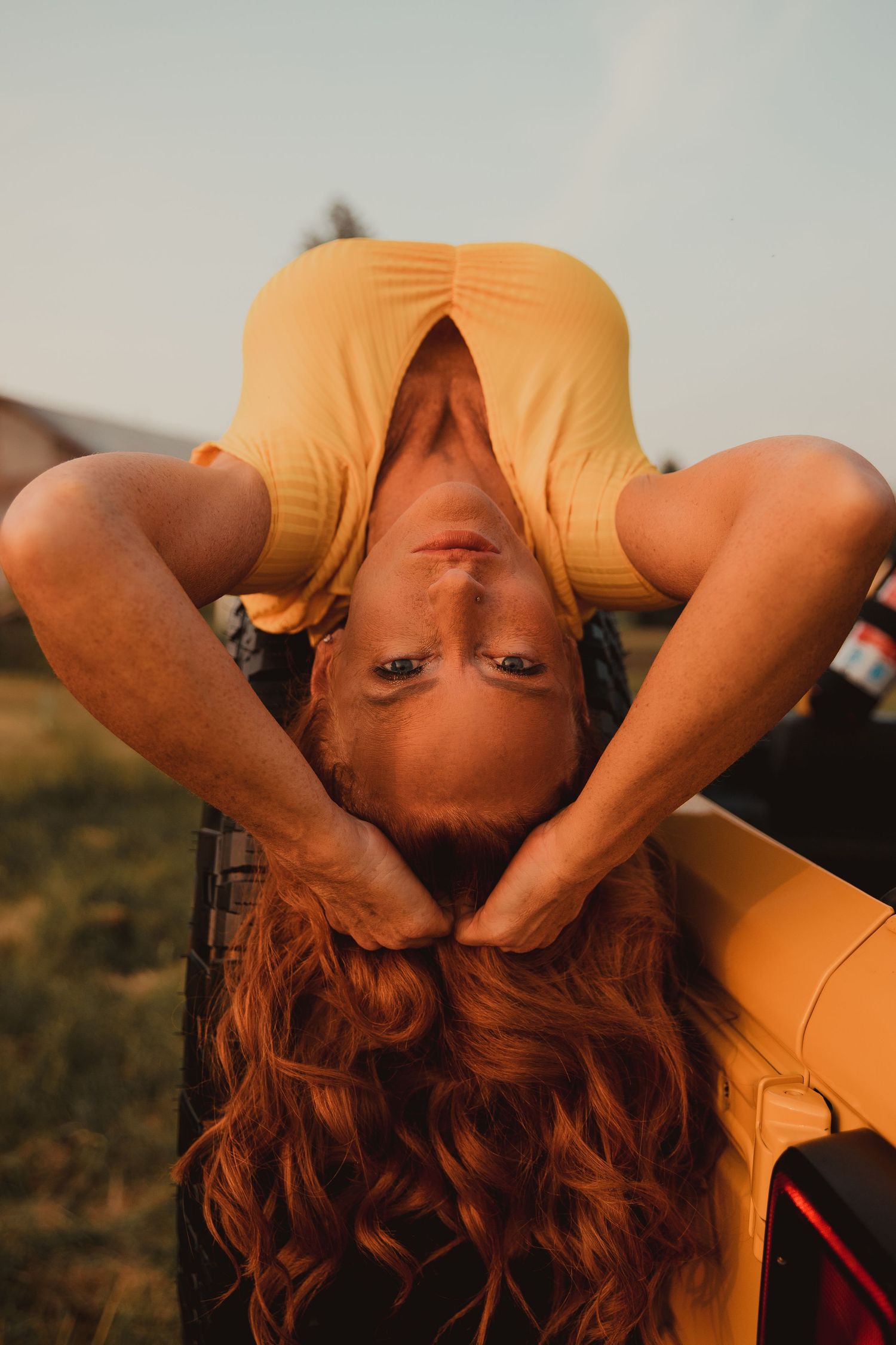 Person with flowing red hair wearing a yellow top poses upside down in golden sunlight outdoors.