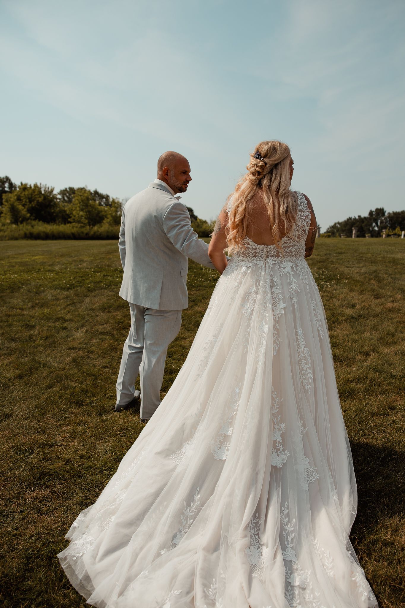A bride in an elegant lace wedding gown with long train stands beside her partner in a white suit on a grassy field.