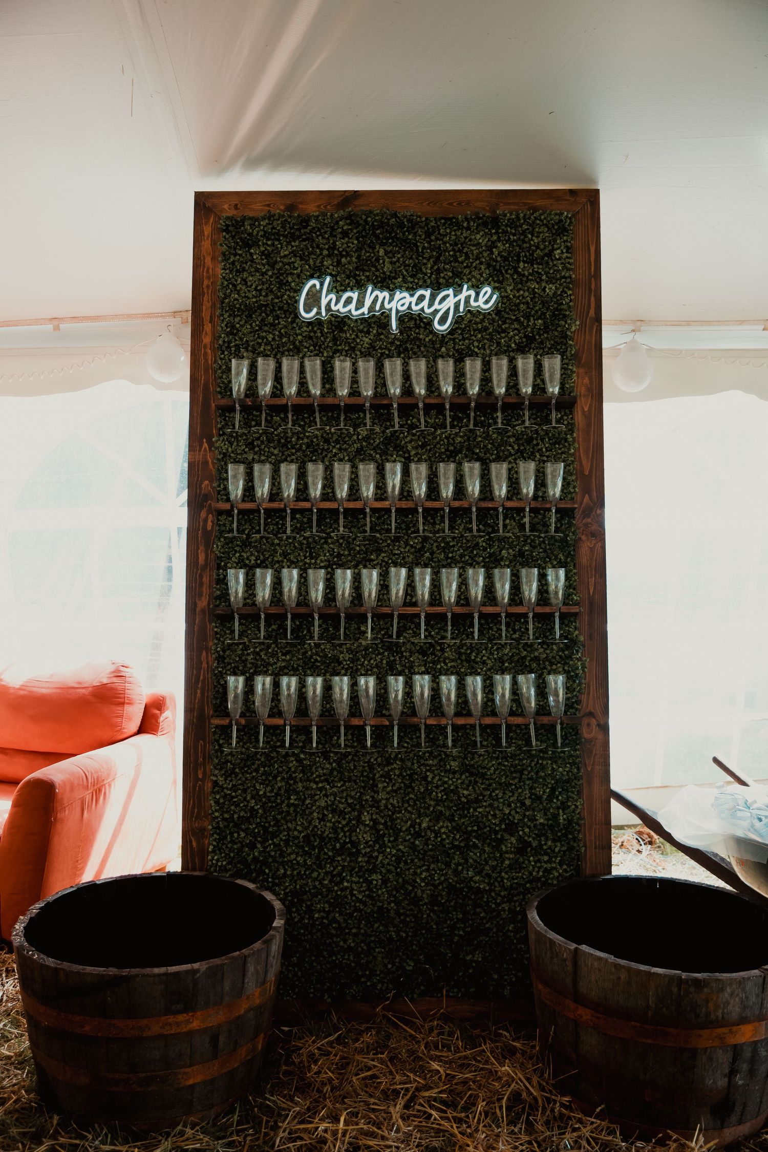 A rustic champagne wall display with rows of glasses mounted on artificial grass backdrop and two wooden barrels below.