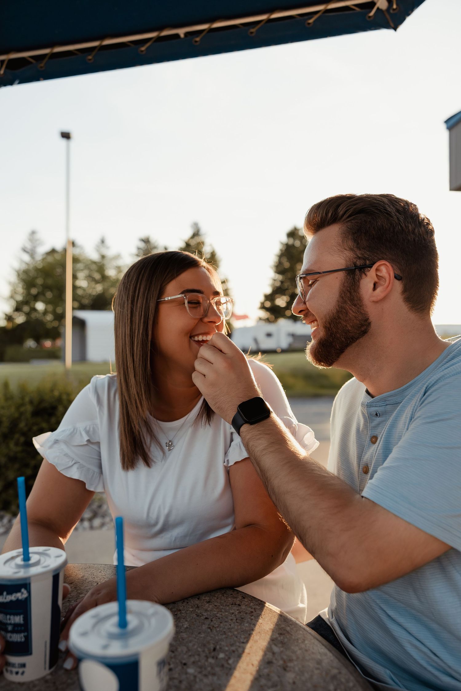 A couple shares a playful moment at Culver's outdoor seating, man feeding woman while both smile and wear glasses.