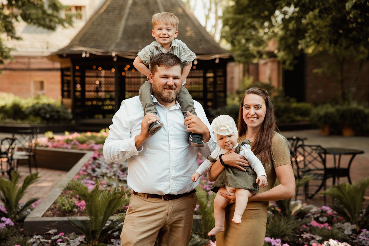 Happy family posing outdoors, father carrying toddler on shoulders, mother holding baby, surrounded by colorful garden flowers.