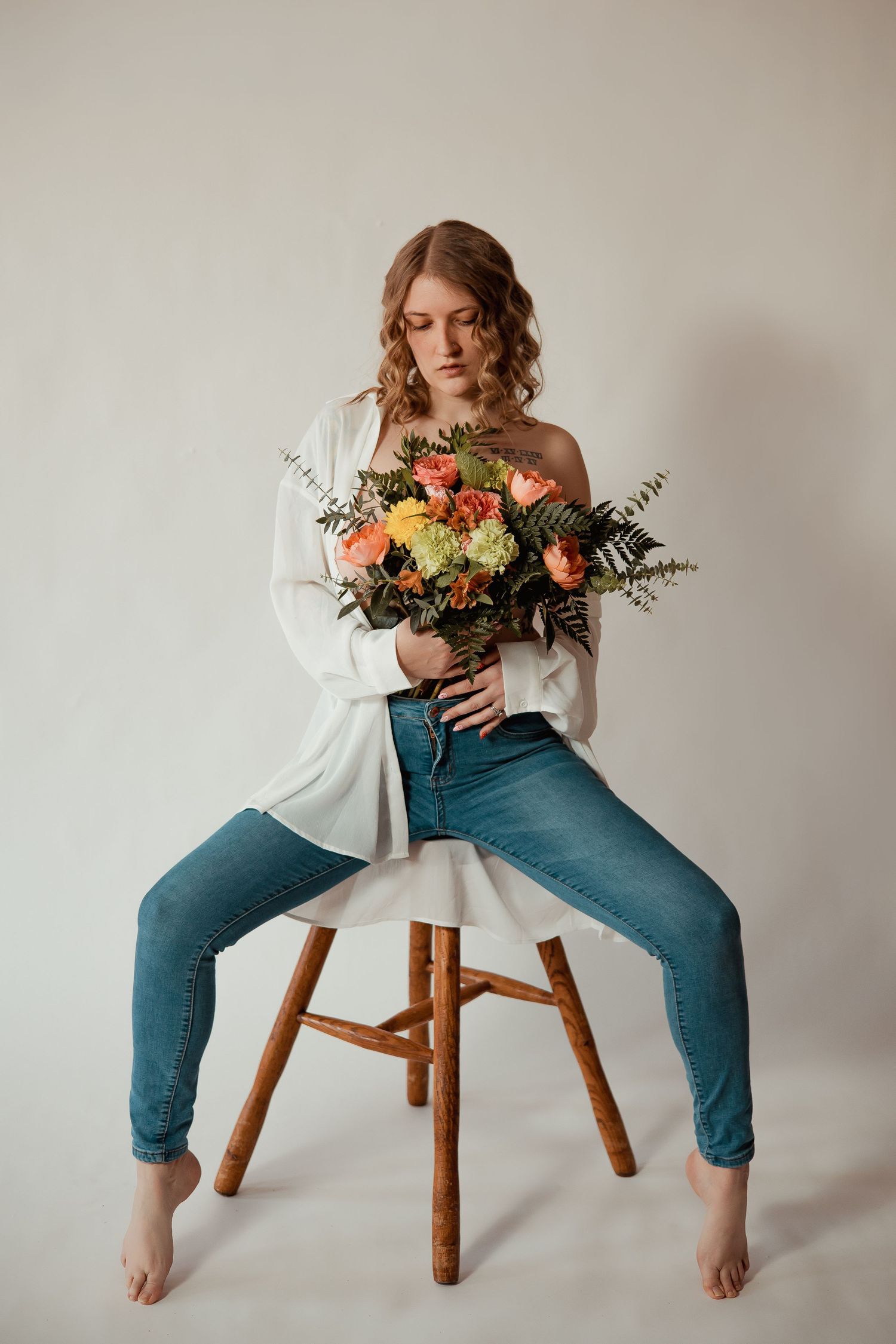 A figure in blue jeans and white top sits on wooden stool holding colorful floral bouquet in artistic poses.