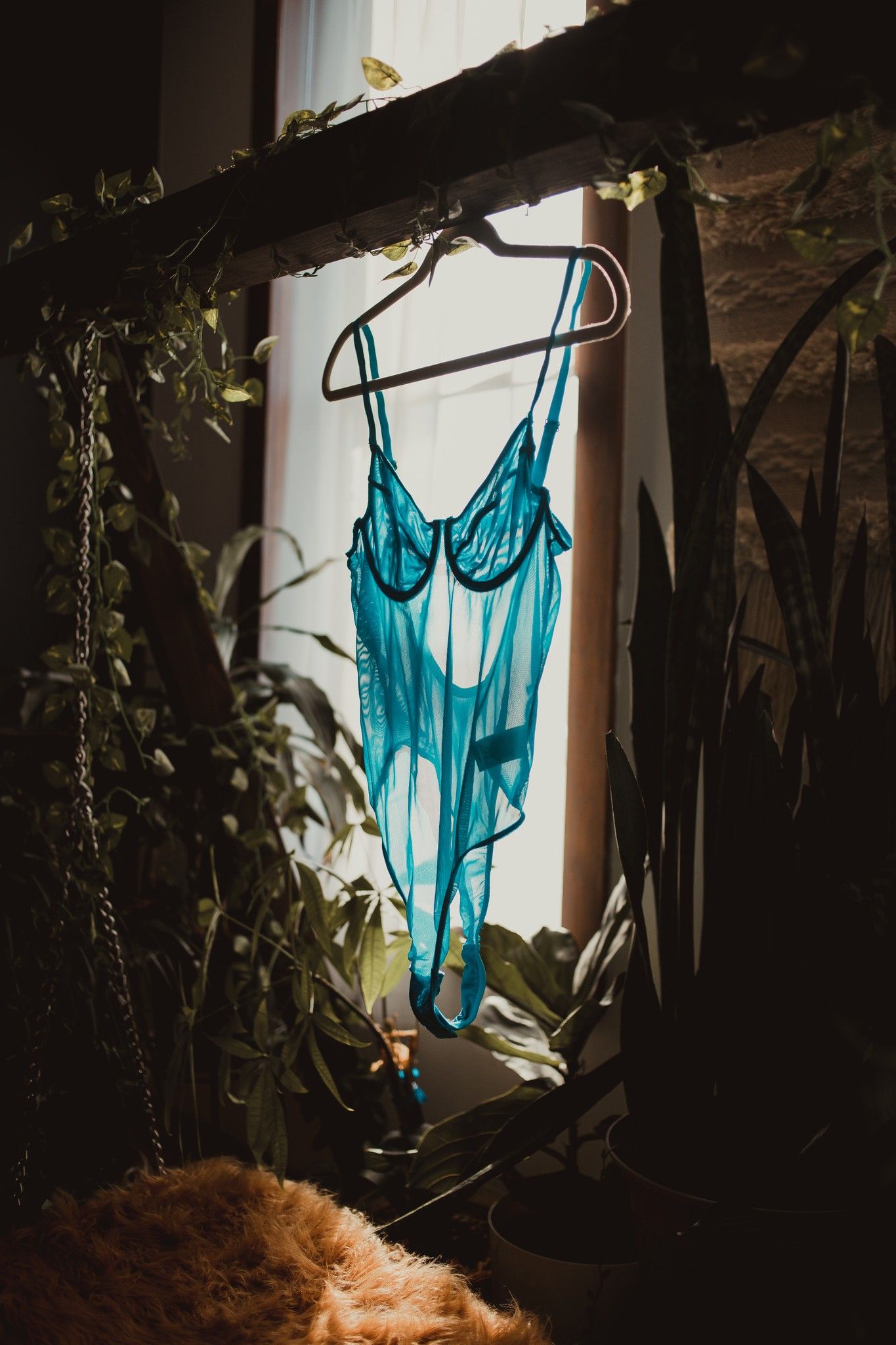 A turquoise lingerie slip dress hangs on a hanger near a window surrounded by green plants and natural sunlight.